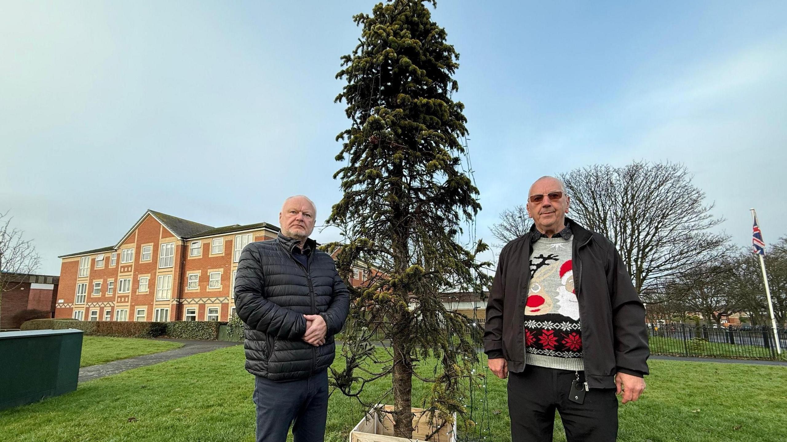 Michael Hartnack and Richard Beck stand next to vandalised Christmas tree which is about 8ft tall and thin-looking. They are on a patch of grass with housing behind.
