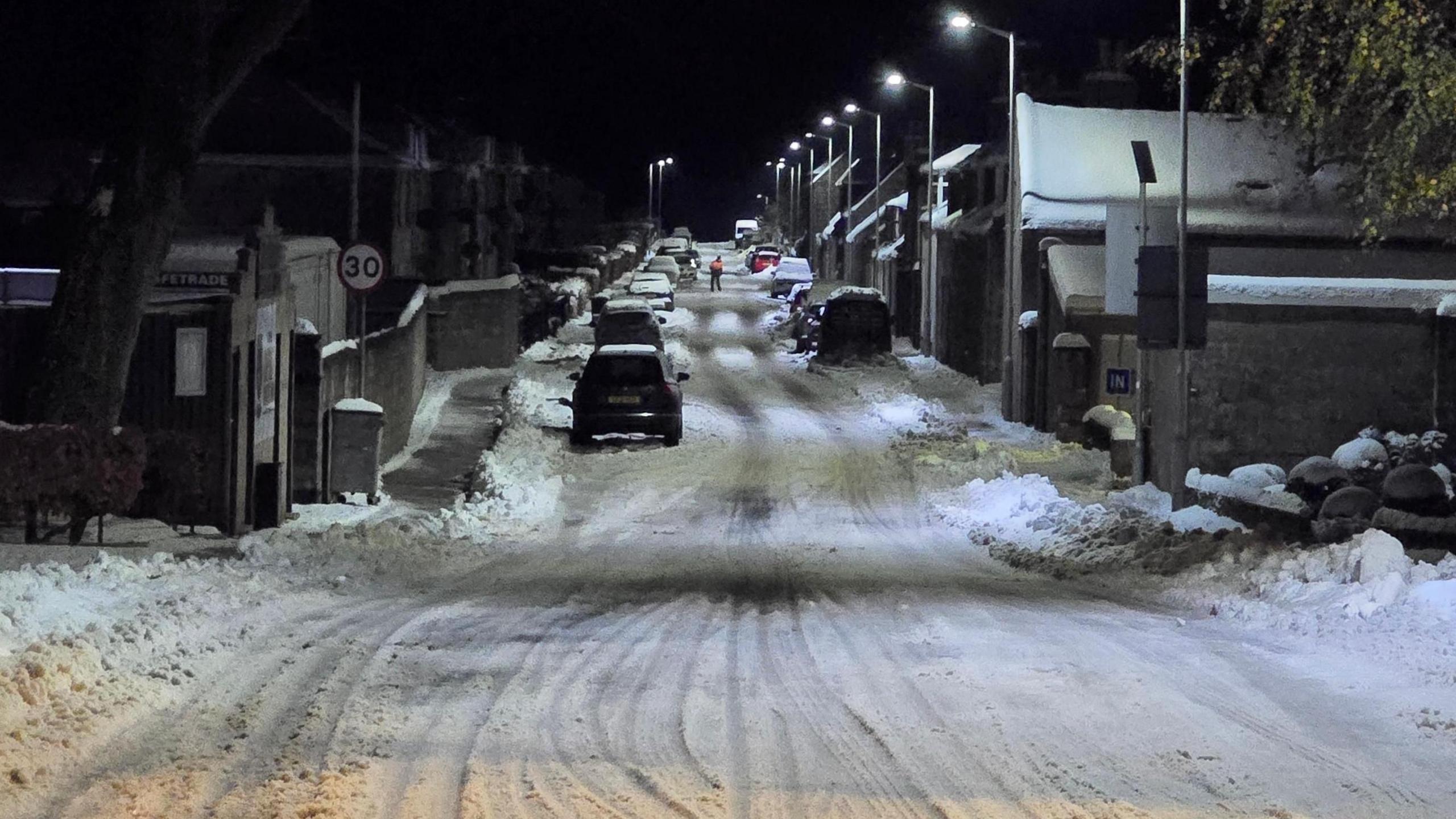 A snow covered road with cars and white streetlights extending into distance.