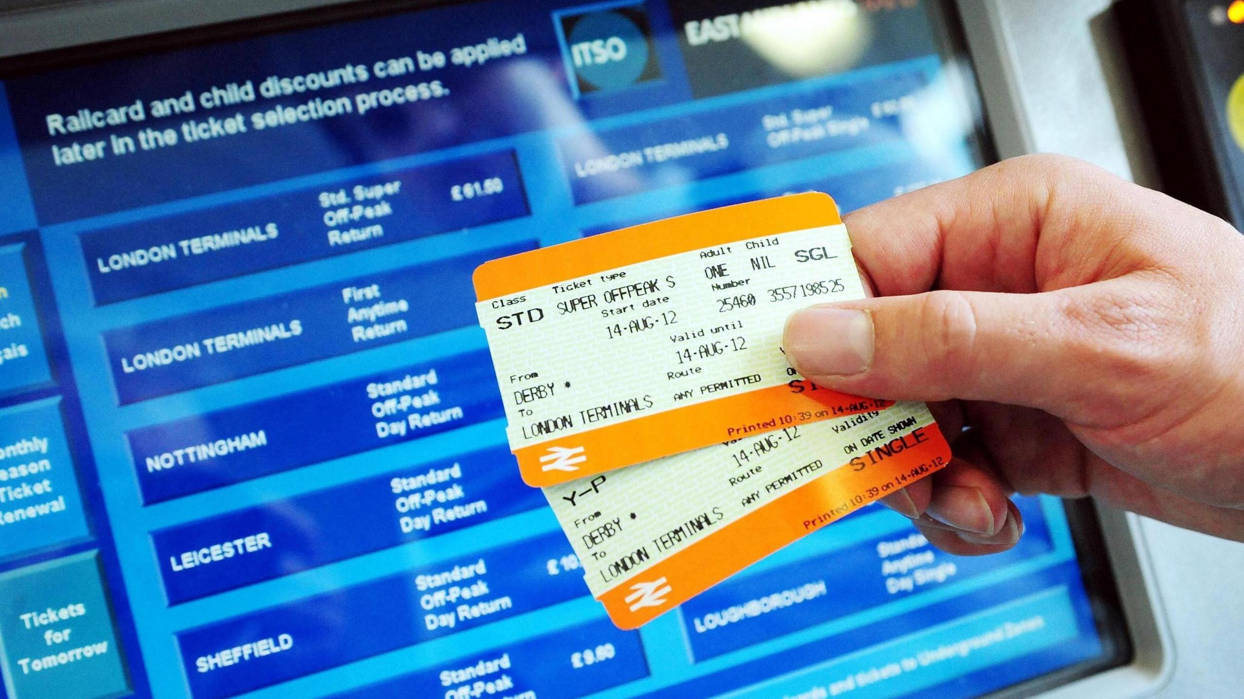 Two kinds of rail tickets are held up in front of a ticket machine displaying a variety of destinations in the UK