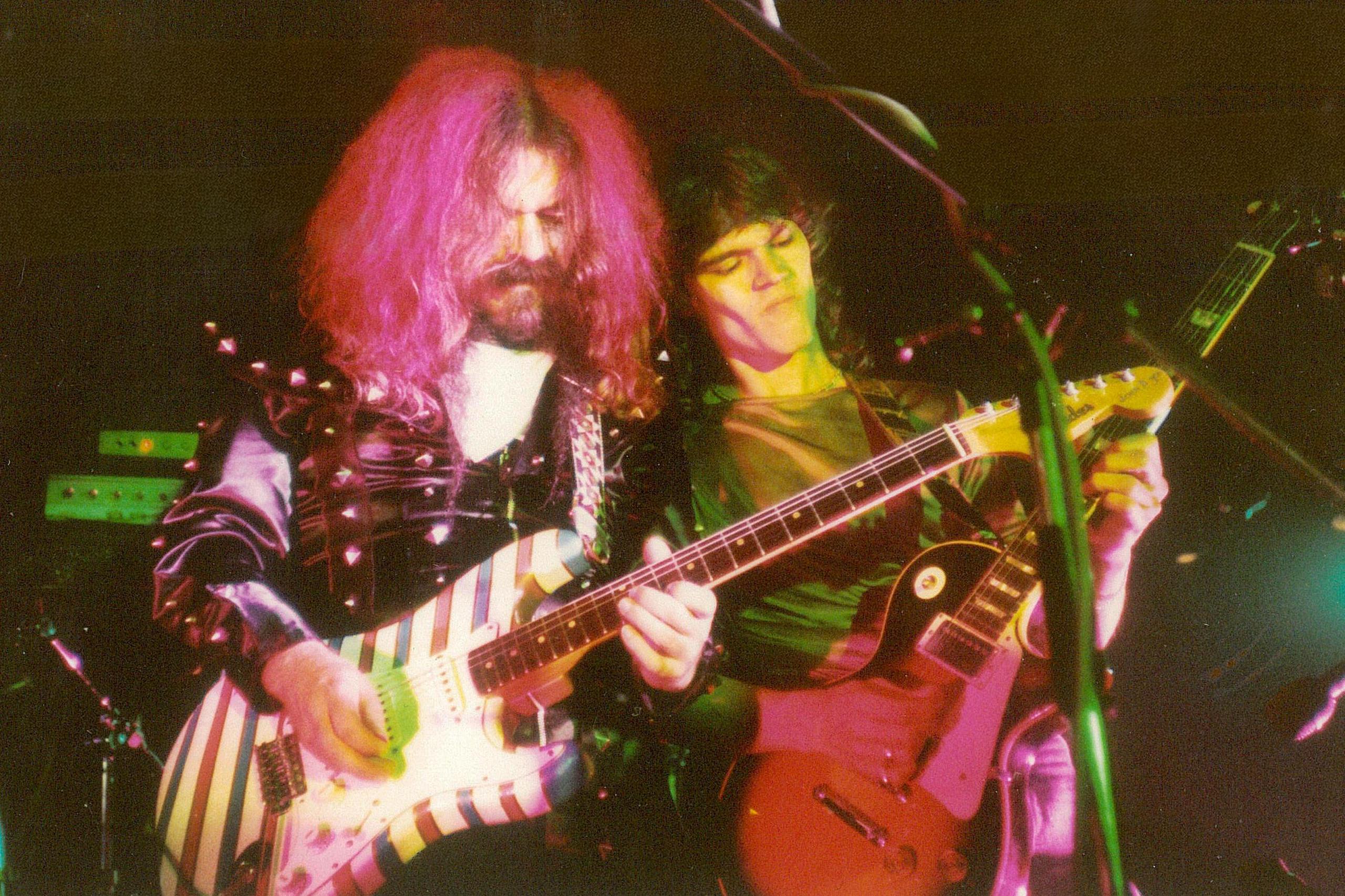 A pink haired Roy Wood is wearing a black satin top and playing a candy-striped guitar. Robin George is beside him also playing guitar. 