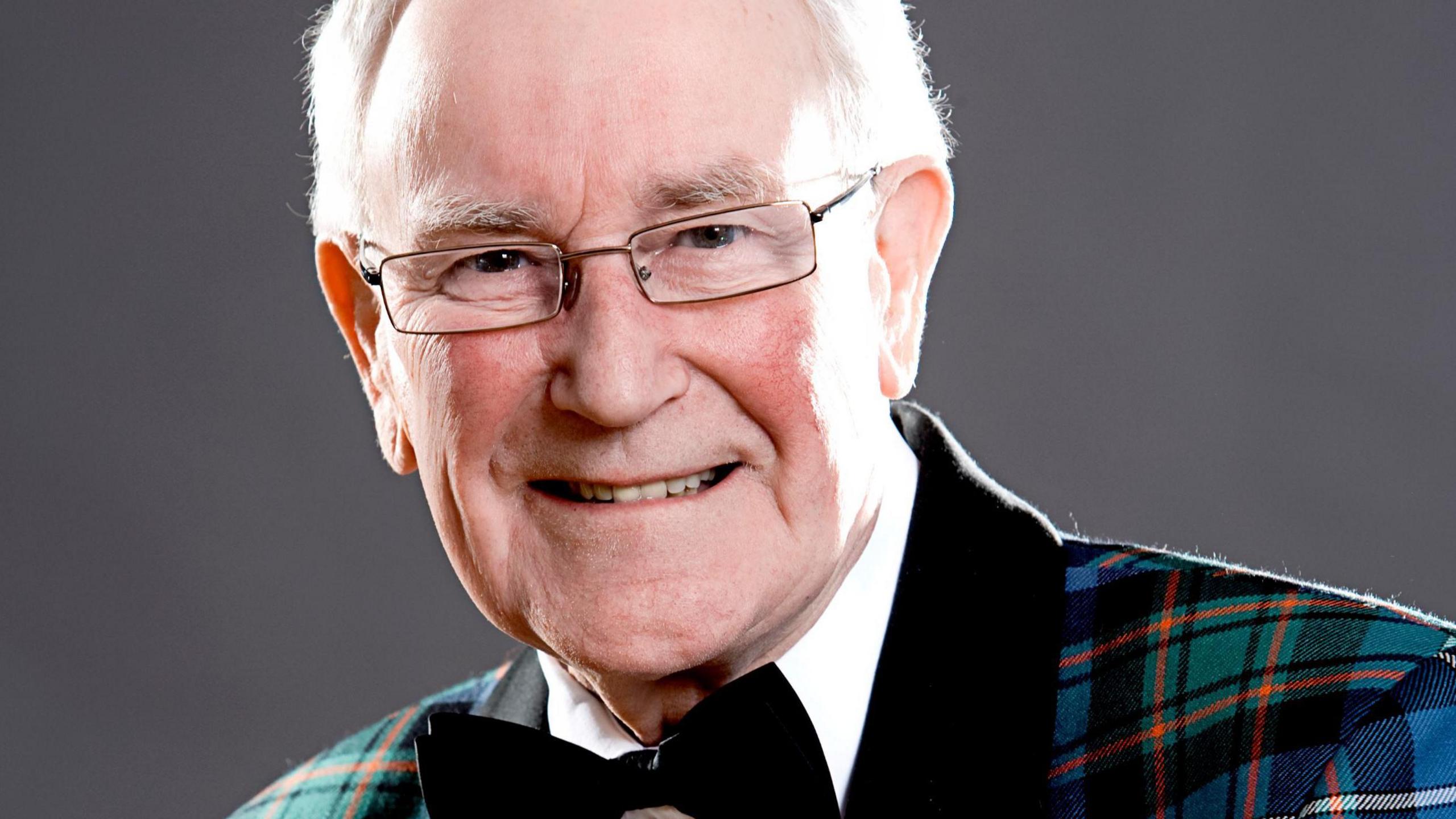 Robbie Shepherd is an older man with white hair. He is smiling and is wearing glasses, and a tartan jacket over a white shirt with a bow tie.