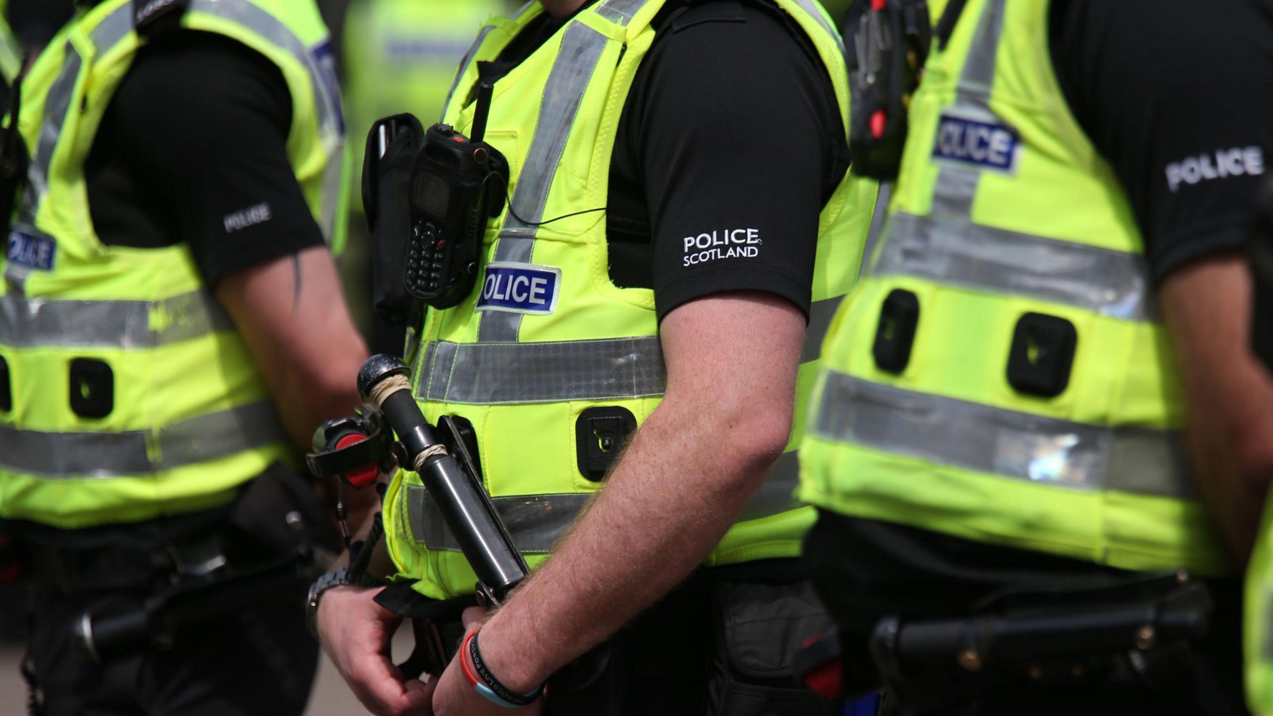 Three Police Scotland officers wearing hi-viz vests over dark clothing standing i a line.