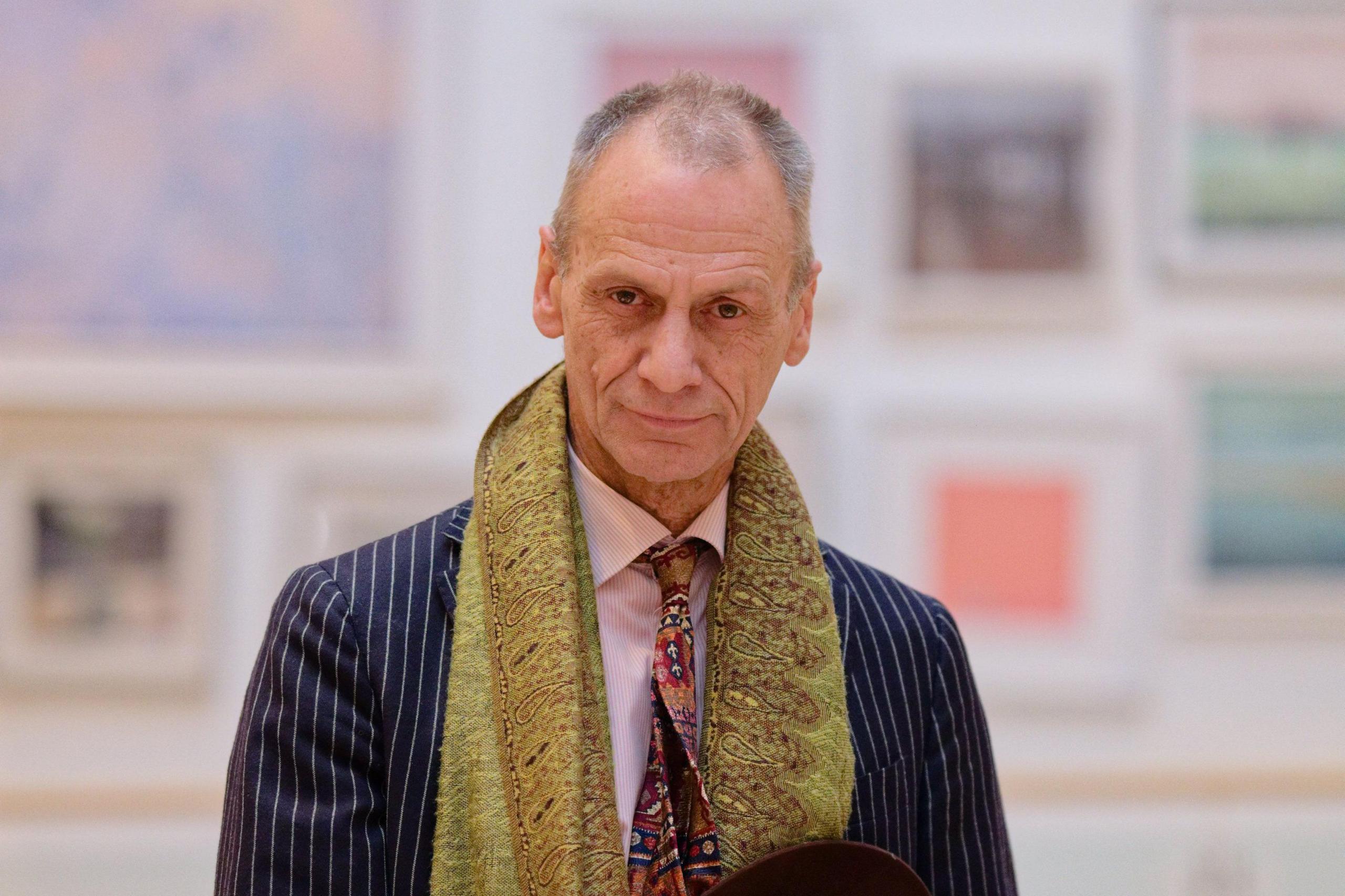 Stephen Powers wears a pin stripe suit and a silk staff. He is looking at the camera and stands in front of paintings