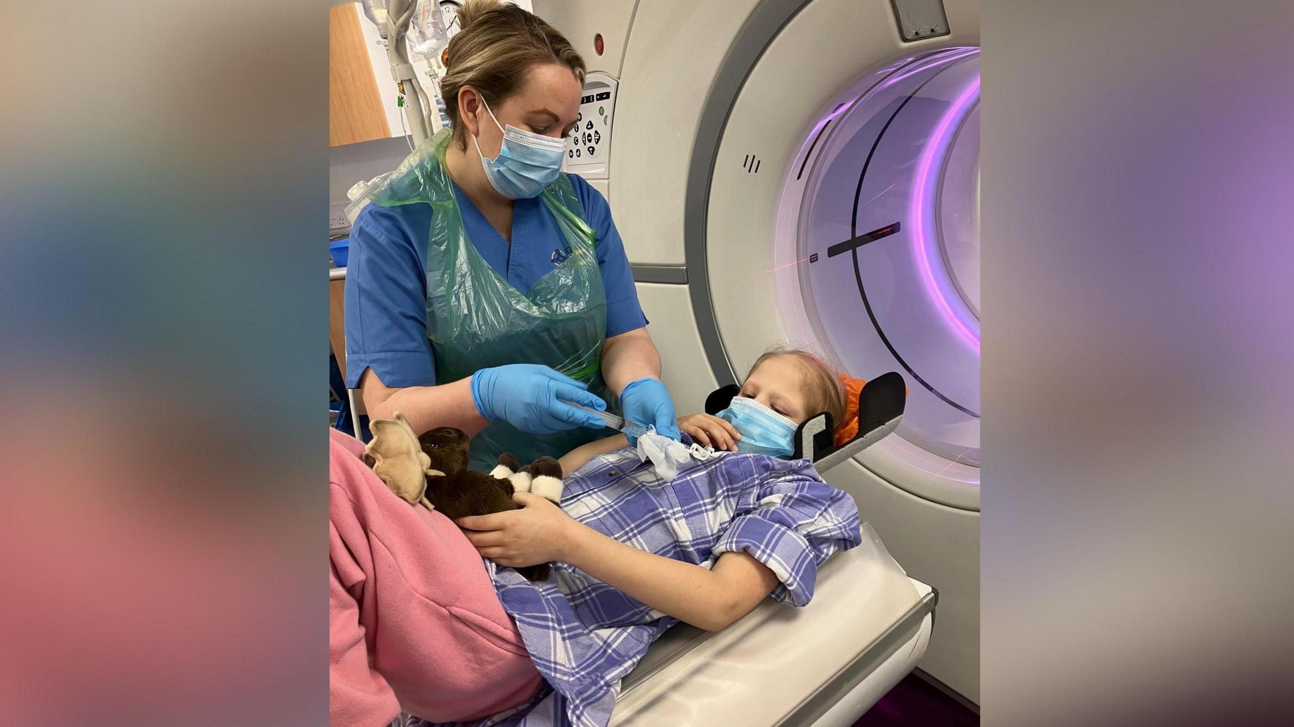 Llun o Katryn yn gorwedd lawr o flaen peiriant CT sgan. Mae'n gwisgo ffrog glas a gwyn ac mae ganddi fasg glas dros ei cheg. Mae nyrs yn sefyll nesa iddi.