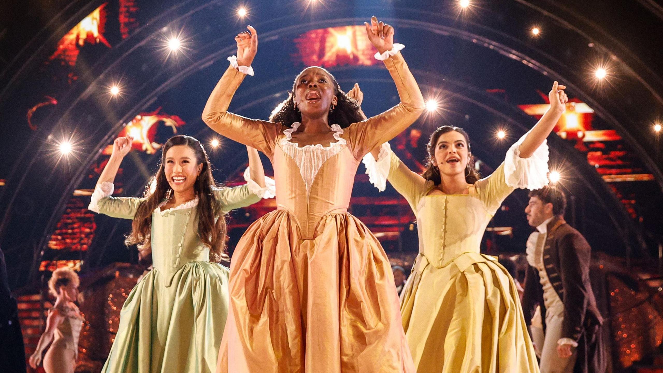 Three female actors and singers dressed as the Schuyler sisters, performing a song on stage at Strictly Come Dancing. There are other actors behind them in period costume.
