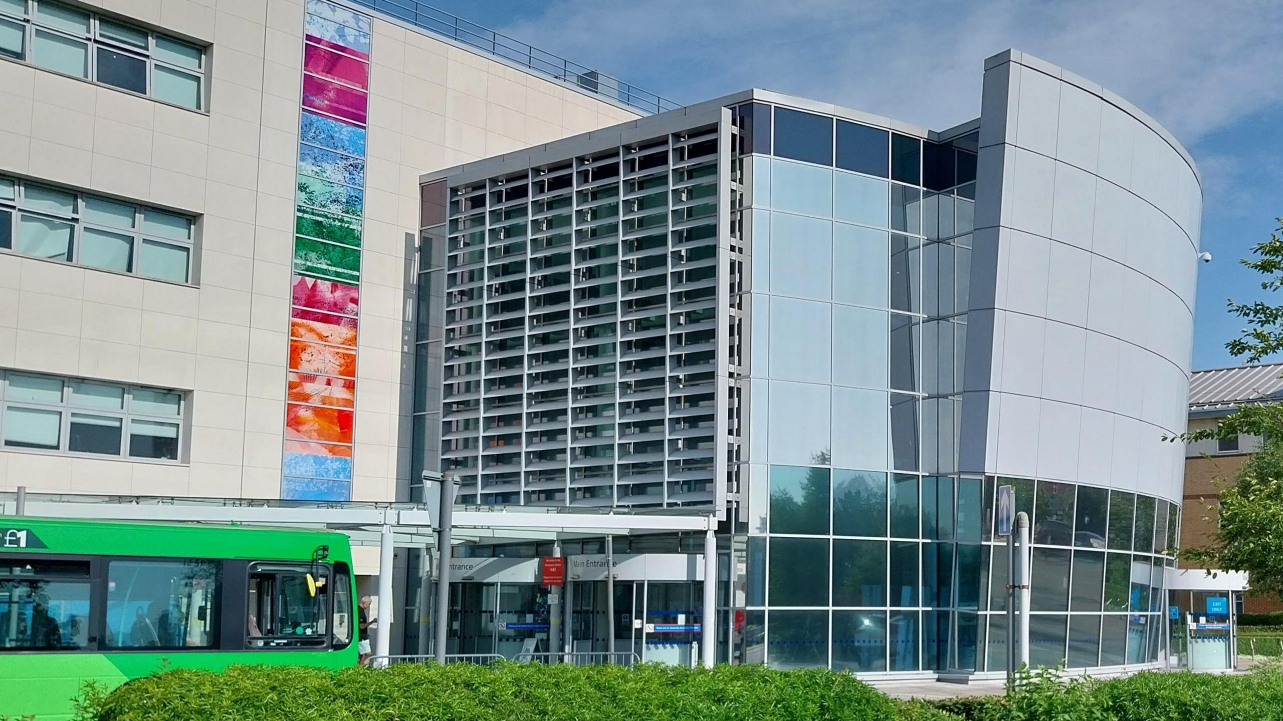 The entrance building of Broomfield Hospital, which is tall and grey with windows. A green bus is passing by in front of the building, with a bush also in view next to the bus.