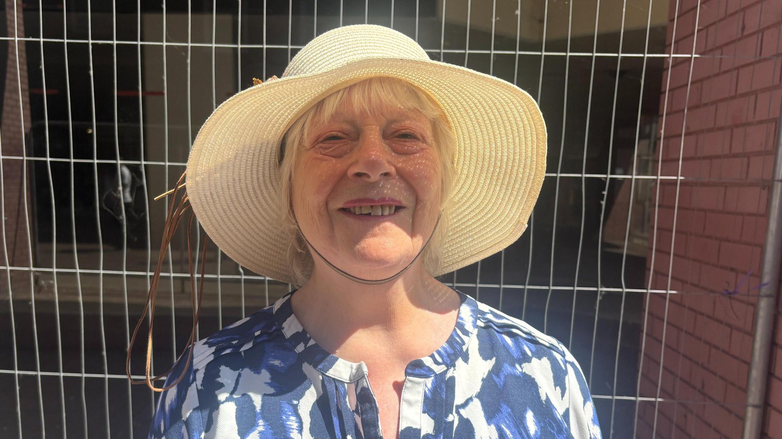 A woman in a sun hat and a flowery top stands in front of Heras fencing.