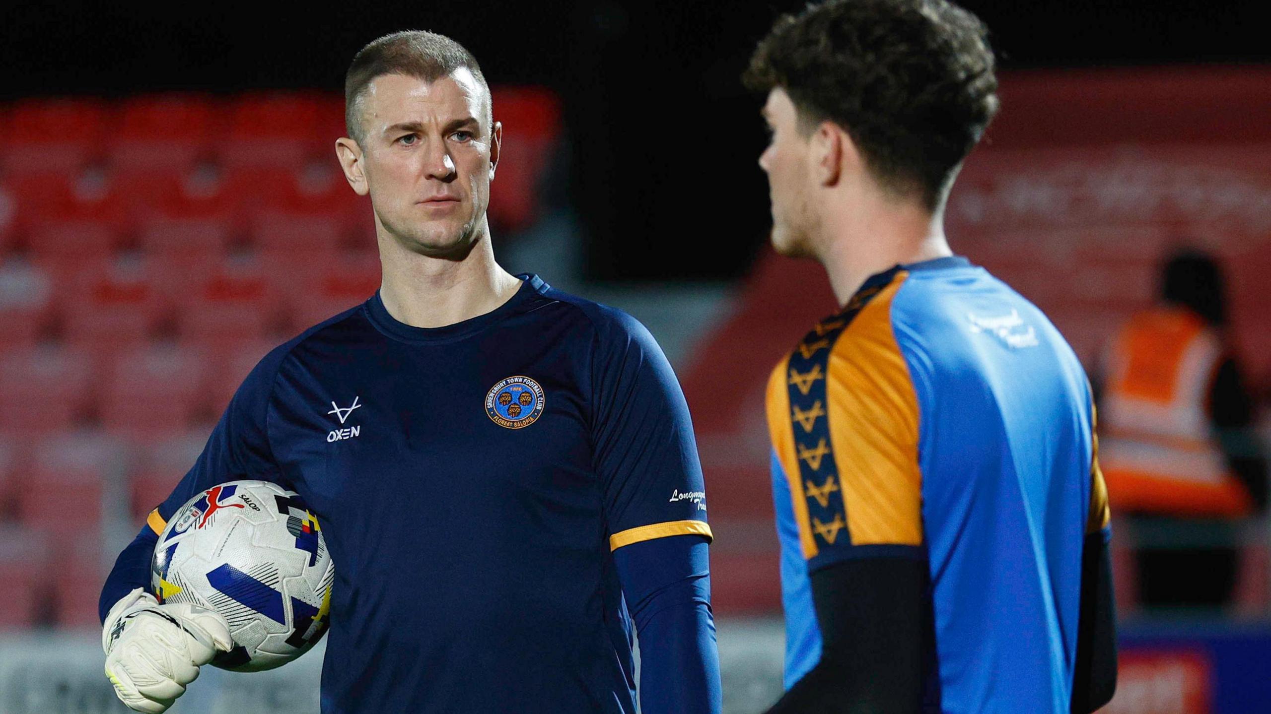 Former England international Joe Hart (left), with gloves on and ball in hand, working as Shrewsbury Town goalkeeper coach for a night and talking to one of the club's players