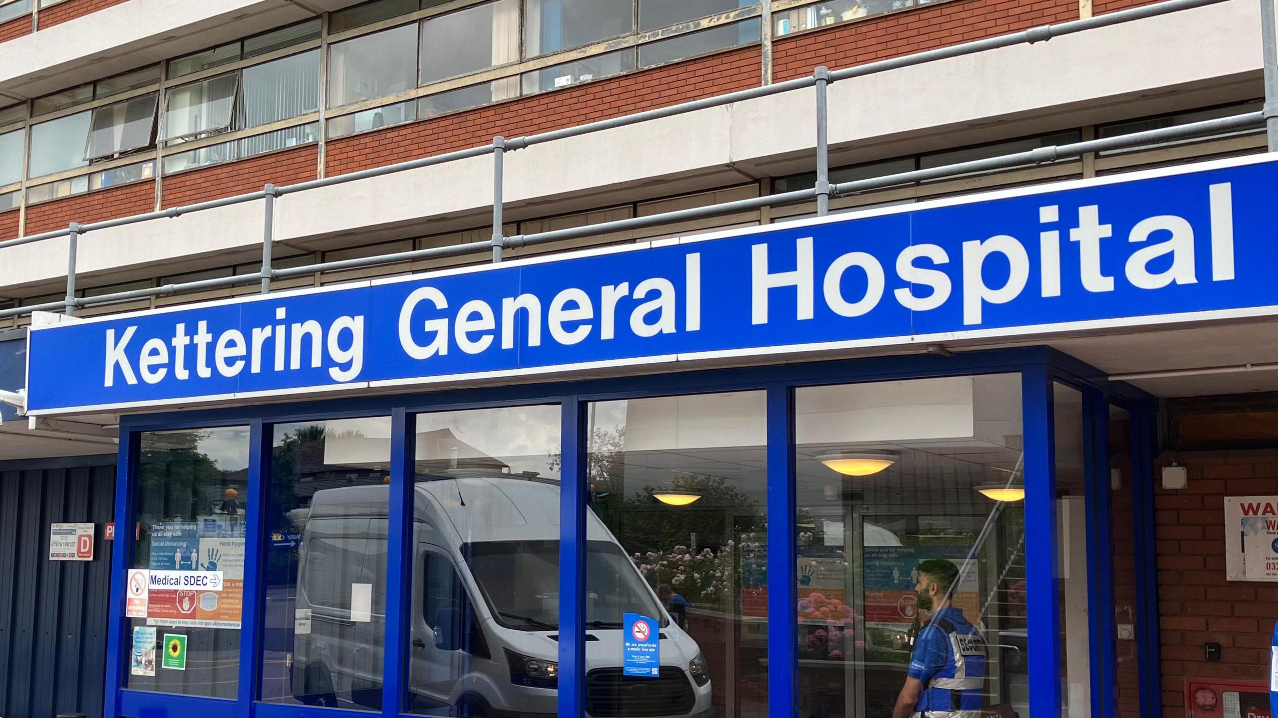 A big blue sign is hanging above one of the entrances to Kettering General Hospital. The hospitals name is written in white, and you can see automatic doors and some of the hospital. 