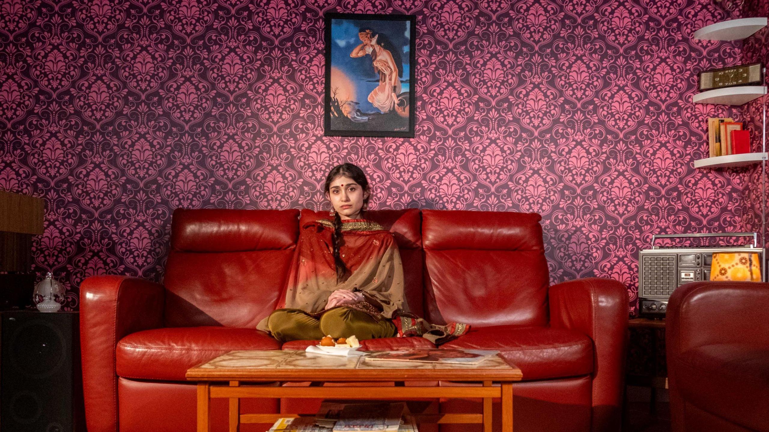 A woman wearing traditional Indian dress with braided hair sits on a red leather sofa with a coffee table in front of her. To her right is a radio and bookshelves and above her head is a piece of artwork of a woman in front of a night sky. The wallpaper is a bright magenta pattern.
