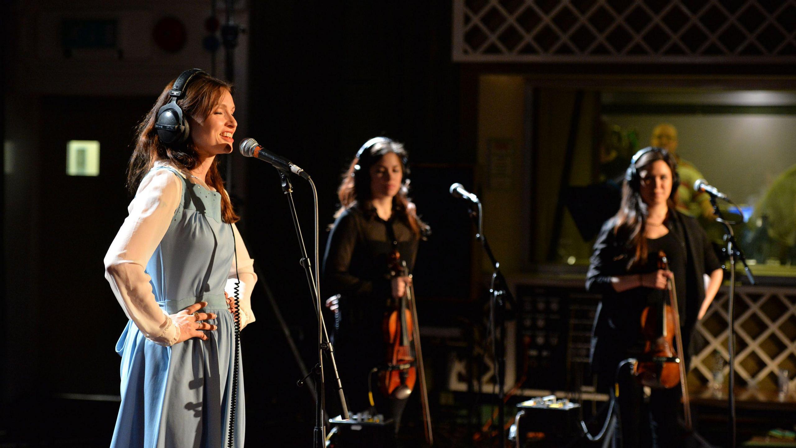 Sophie Ellis Bextor is singing into a microphone at a recording studio. She has her hands on her hips and eyes closed. She has long brown hair and is wearing headphones and blue dress with white sleeves. Next to her are two female backing singers.