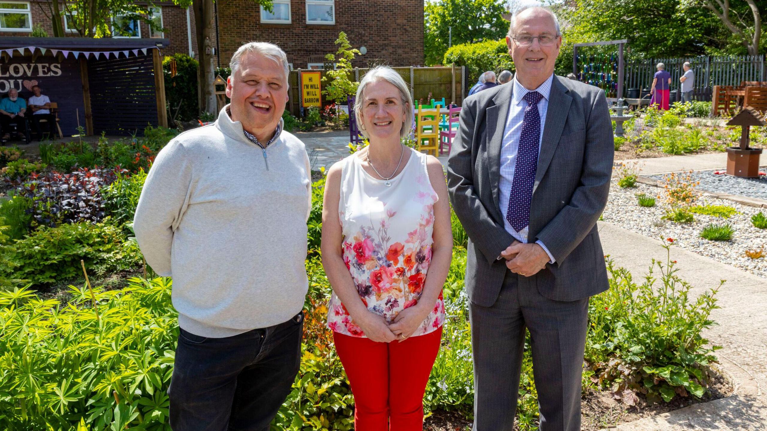 Two men and a woman stand in front of a garden featuring plants, pathways and children's furniture.