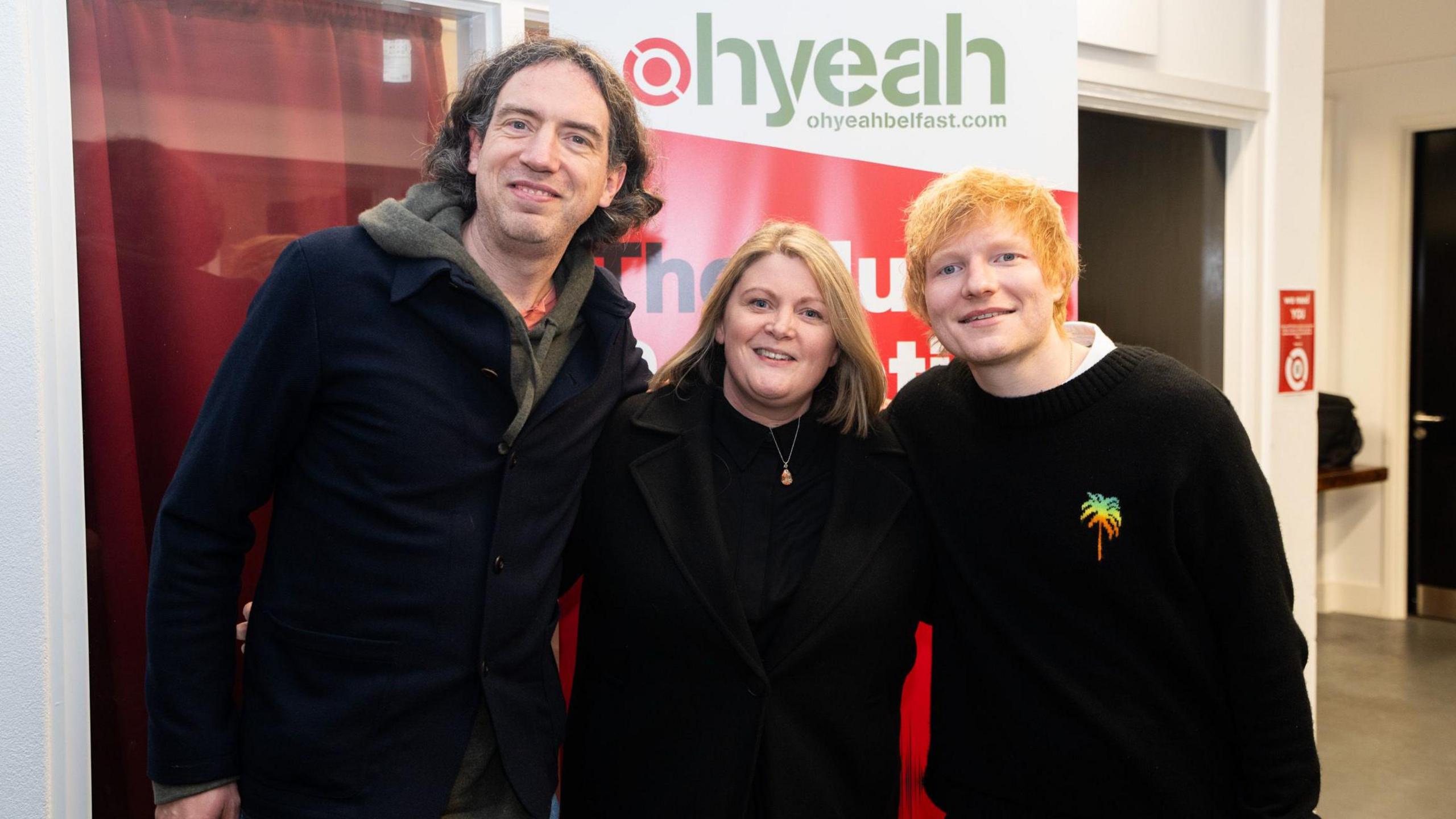 Gary Lightbody and Ed Sheeran at the Oh Yeah Music centre. Gary wears a khaki hoody, with a navy jacket over it and jeans. He has curly longish hair. Ed Sheeran has shaggy red hair and wears a black jumper with a colourful pineapple logo on it. They are standing on either side of a woman in a long black coat, black jumper and acorn necklace.