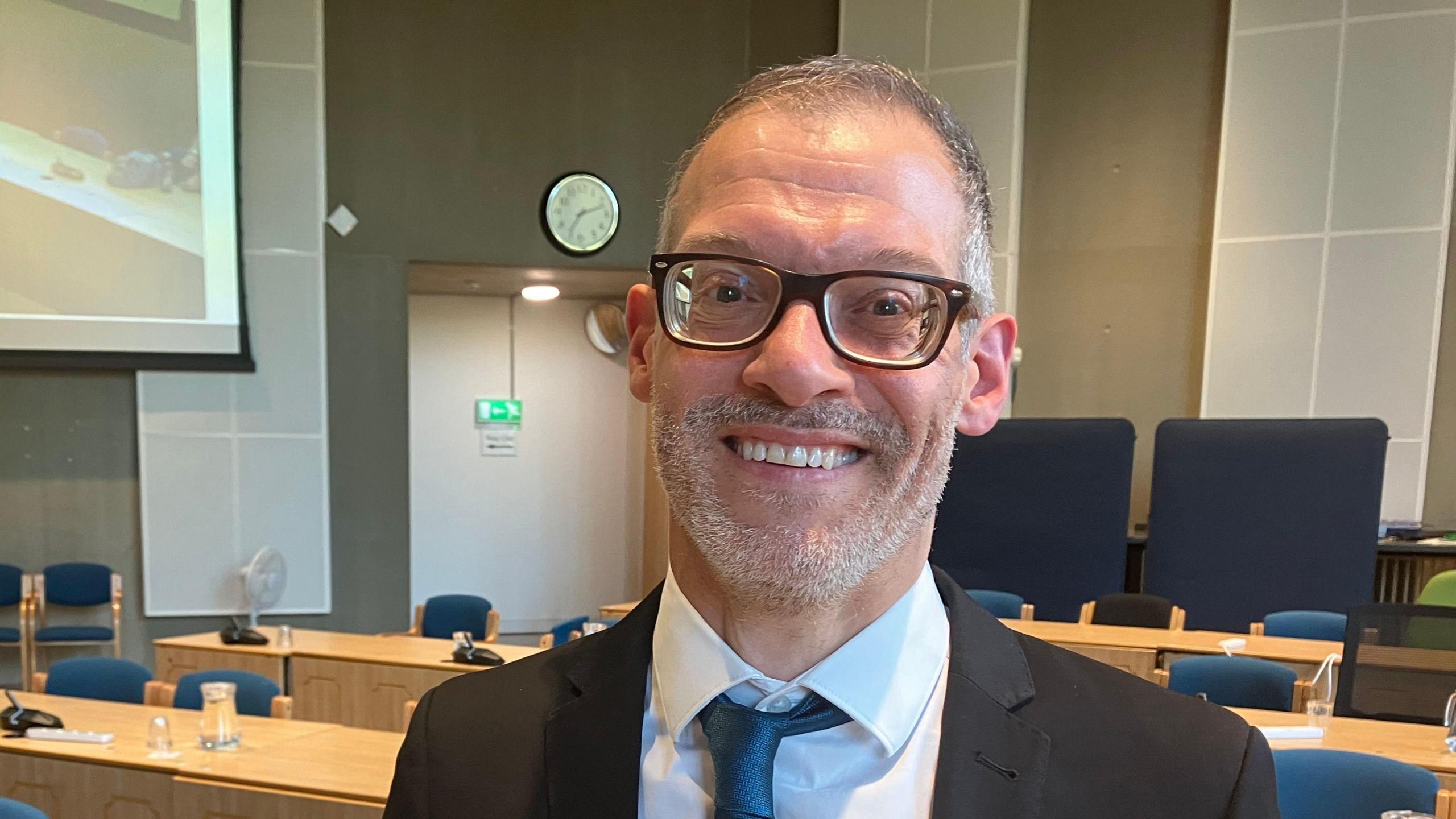 Central Bedfordshire Council Independent Leader Adam Zerny smiles ayt the camera. He is wearing a black suit with a dark blue tie and white shirt. He has grey hair and beard and is standing in an empty council chamber.