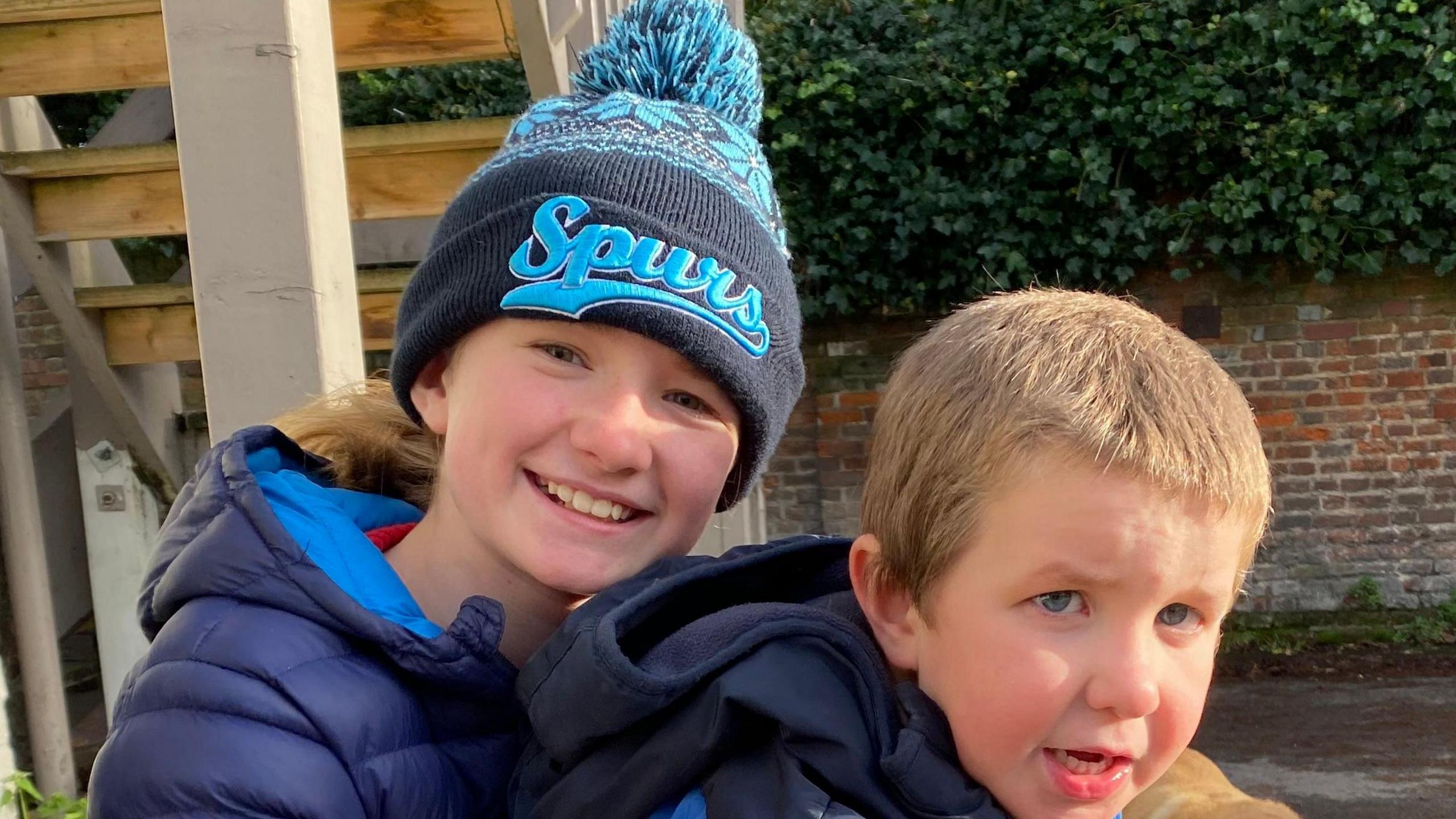 Alex is smiling for the camera while holding Teddy on her lap. She is wearing a blue "Spurs" hat and wearing a blue coat. Teddy has a blue fleecy coat and has short mousey brown hair.