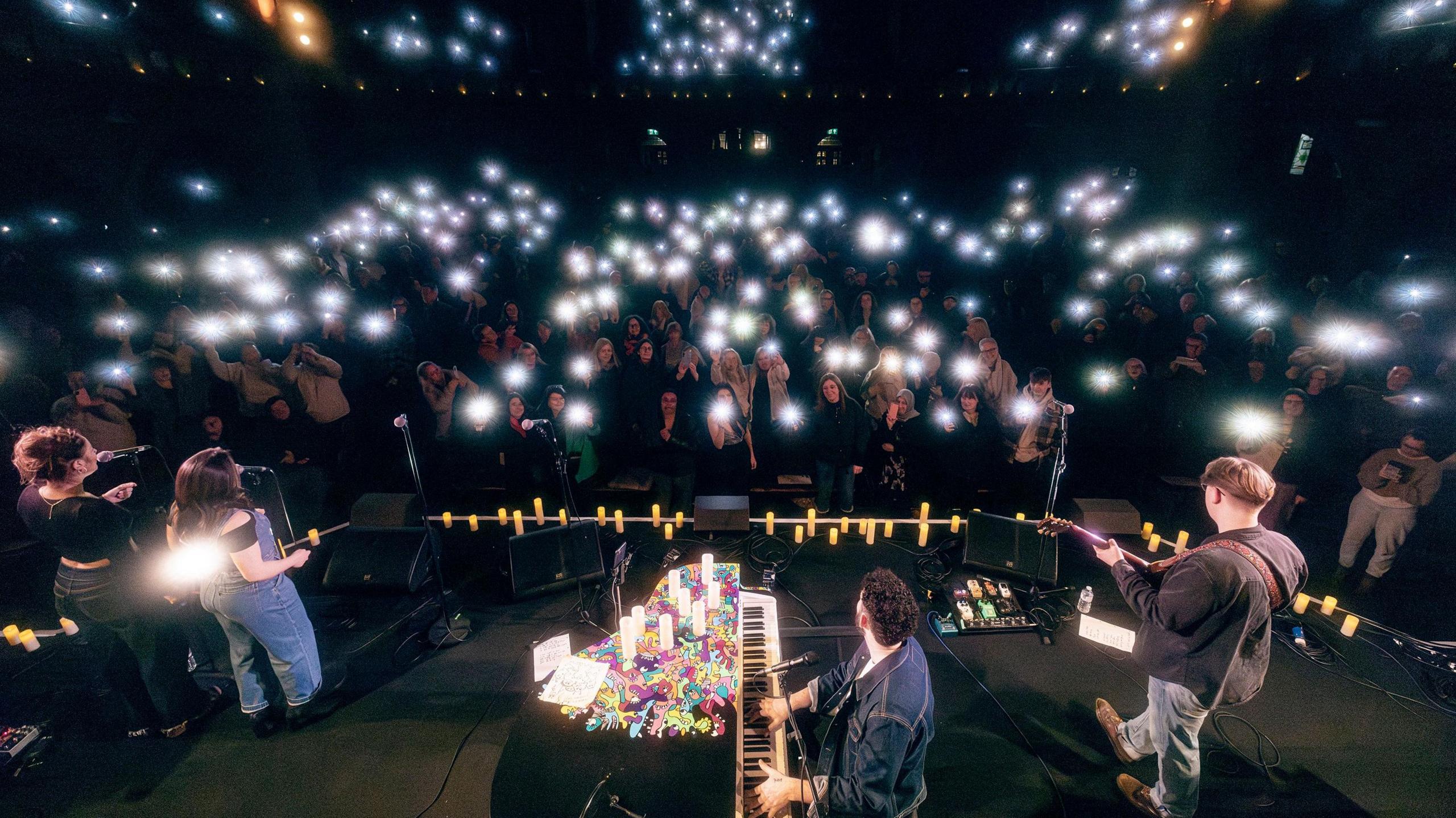 Billy Lockett is sitting at a piano at the front of the picture on a stage looking out at an audience. The piano is covered in flowers and candles. Also on the stage is a guitarist and two singers. The room is largely dark and in front of the stage a large crowd is watching, the majority holding up a mobile phone with the torch on, creating a sea of lights.