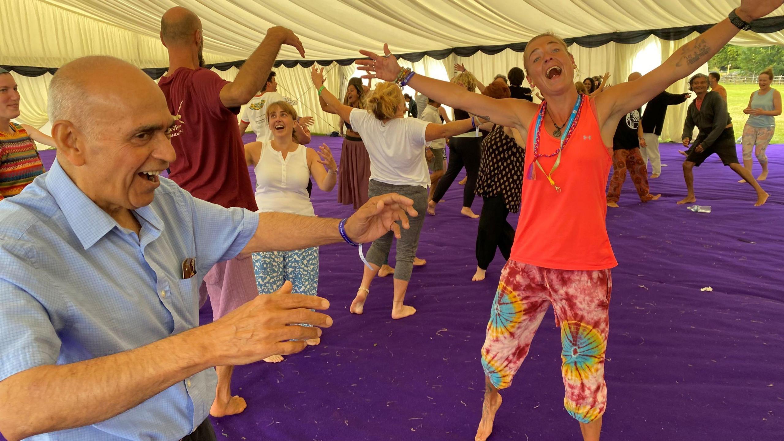 People in a festival tent holding up their arms and smiling and laughing. There is a woman near the front in red with her arms wide apart and her mouth open, as well as an older man in a blue shirt, doing similar.