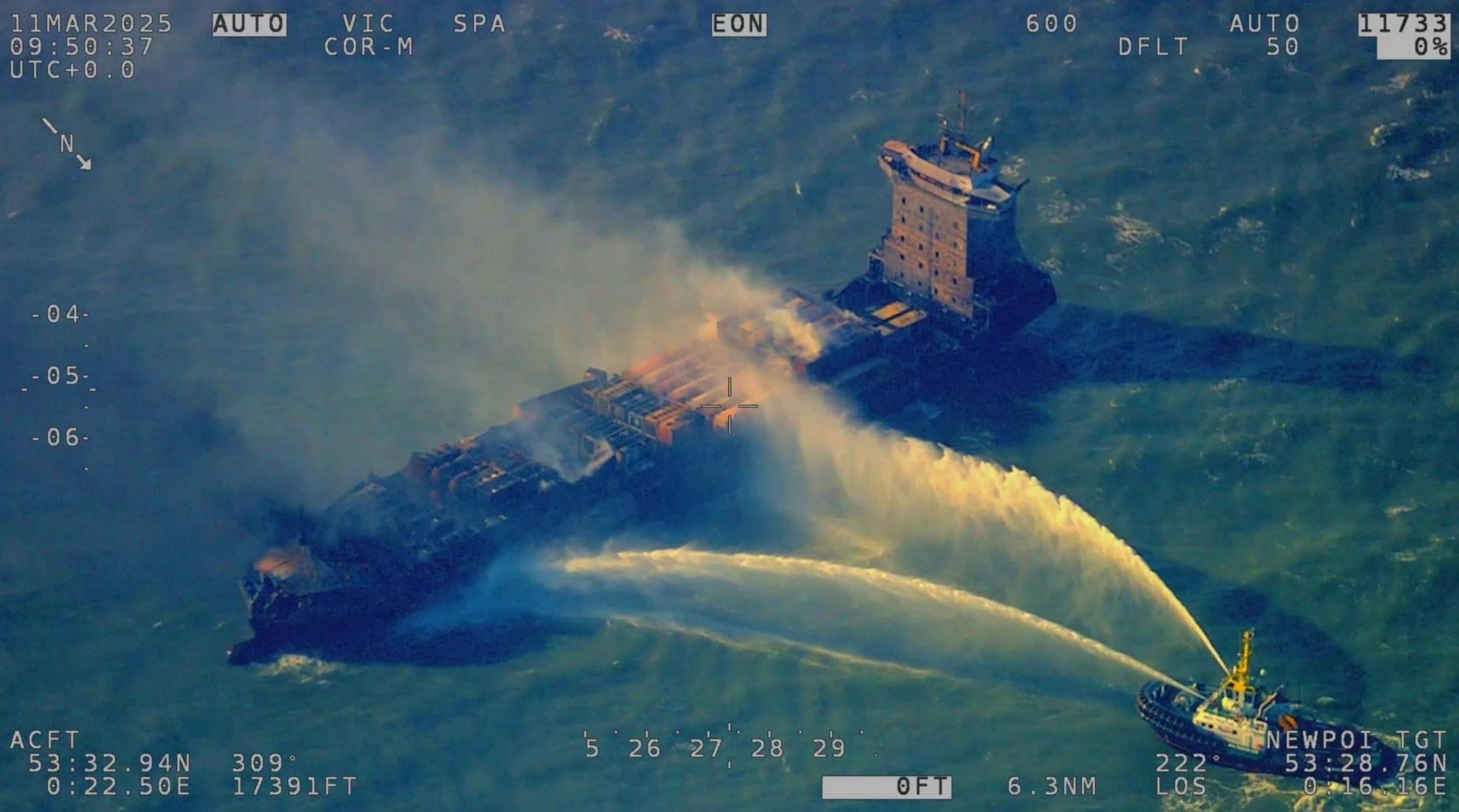 Aerial view of the Solong being sprayed with water from another boat. Flames, smoke and fire damage can be seen. The sea is a deep blue colour.