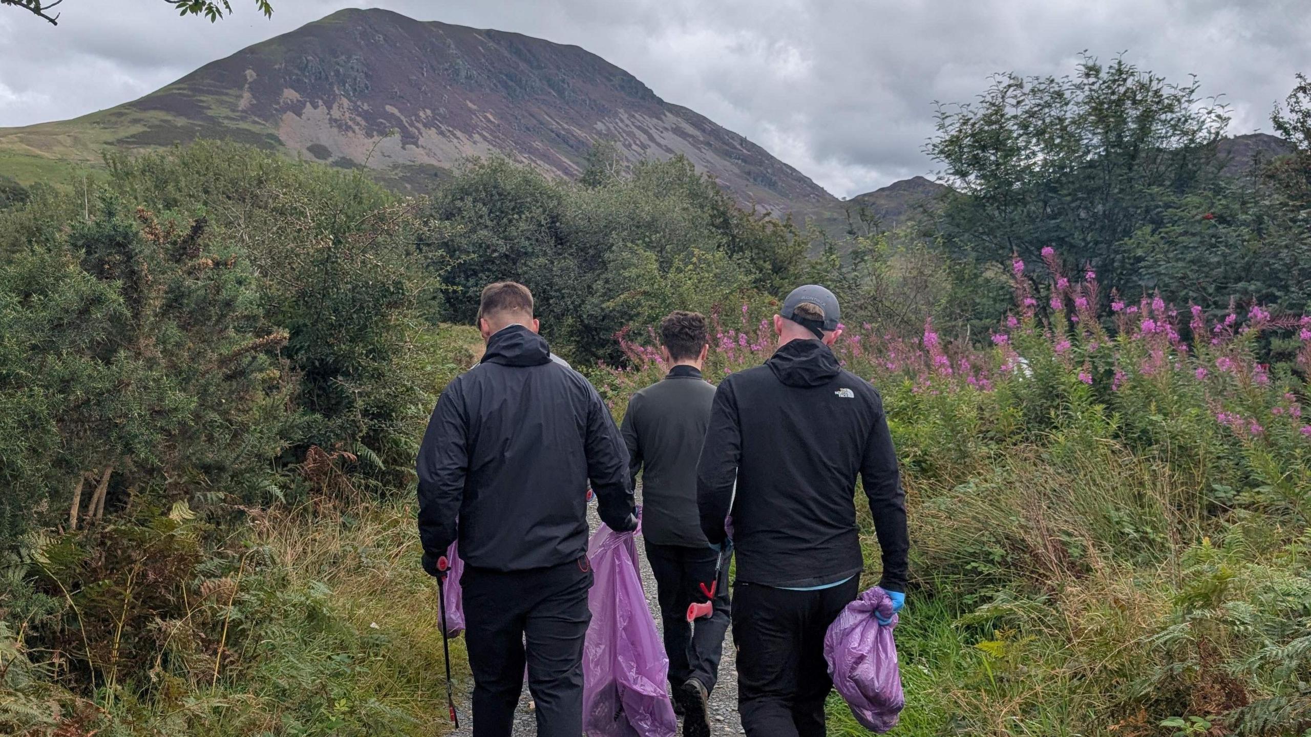 Three young men in dark clothing pictured from the back walking down a path at Ennerdale. They are carrying purple bin bags.