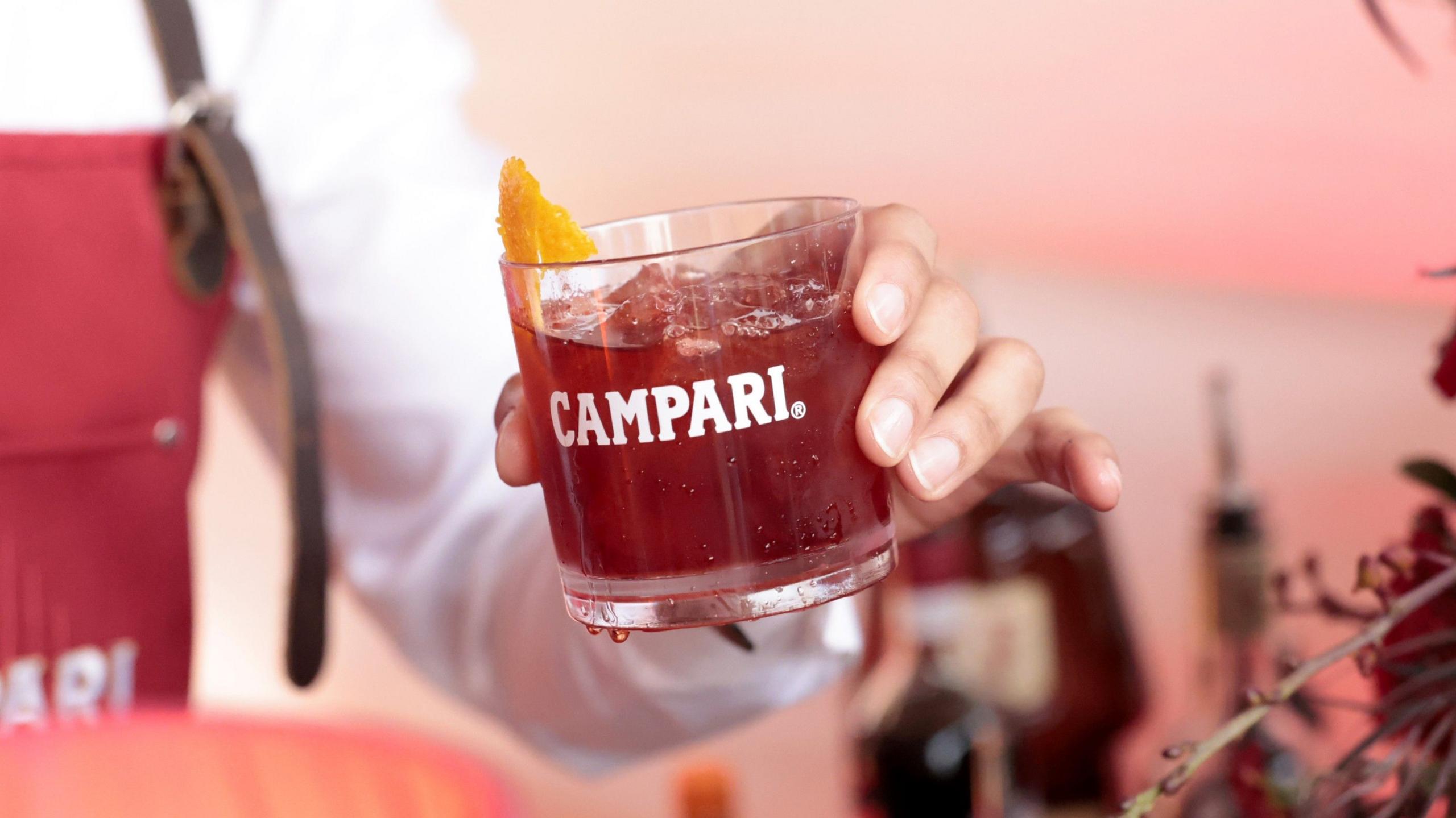 A bartender holds a branded glass of iced Campari with a twist of orange peel.