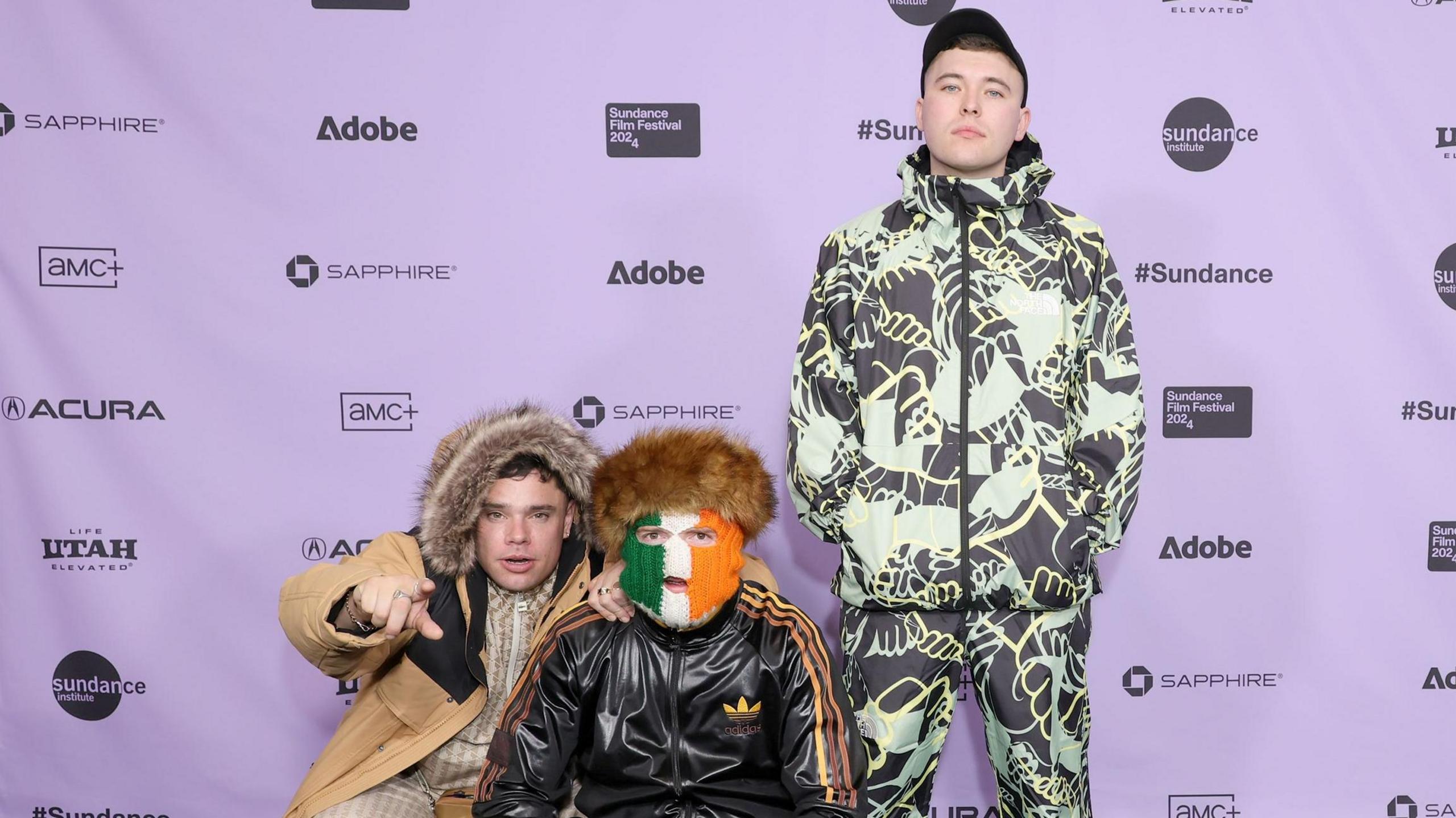 Three young men are standing on front of a purple backdrop. Two are leaning down to pose. One is standing. The man in the middle is wearing a green, white and orange balaclava. 