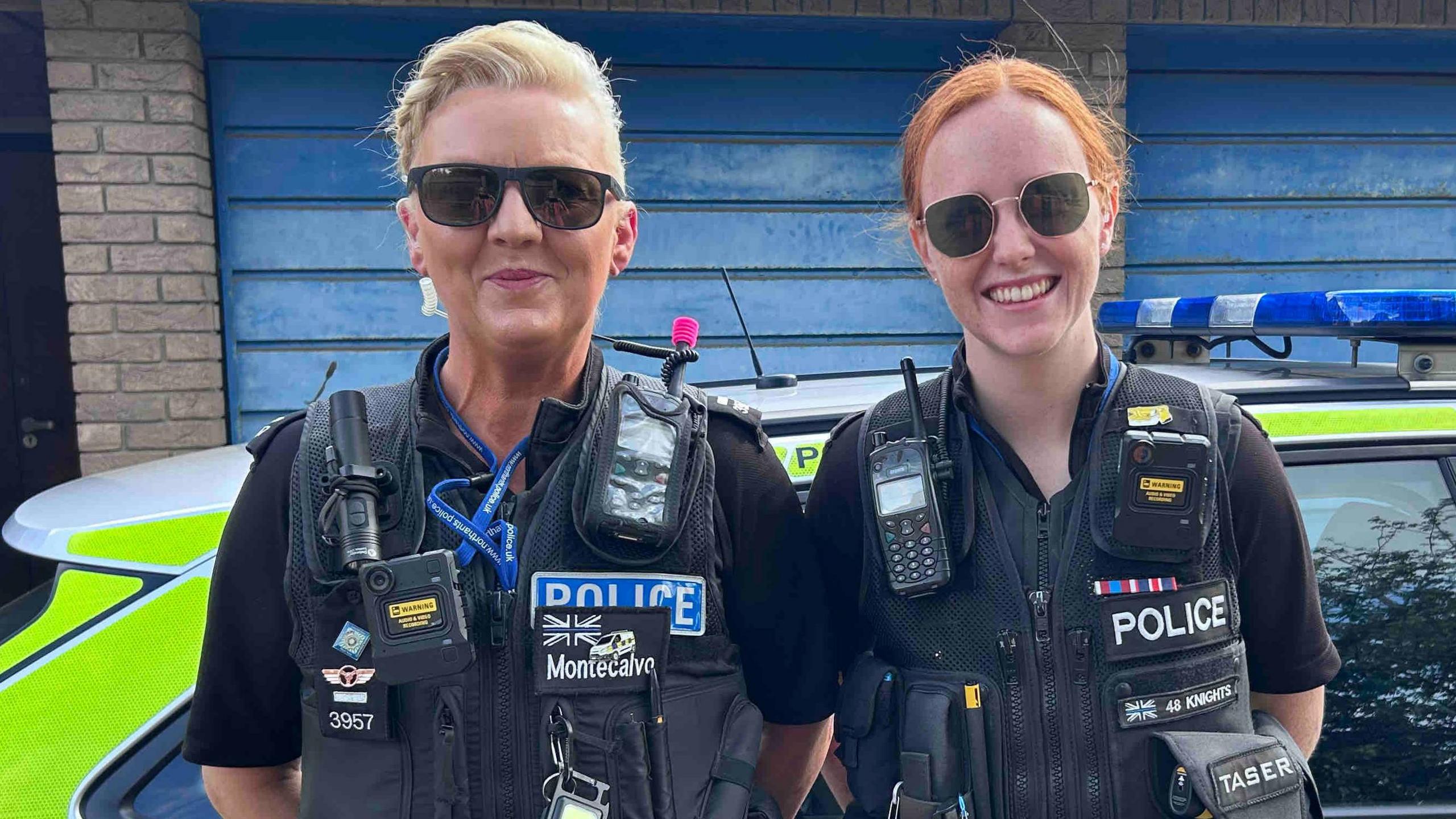 On the left is Lorna Montecalvo who is standing next to PC Annabel Knights. Both women are stood in front of a marked yellow and blue fluorescent police car. They are both wearing police uniform and sunglasses over there eyes.