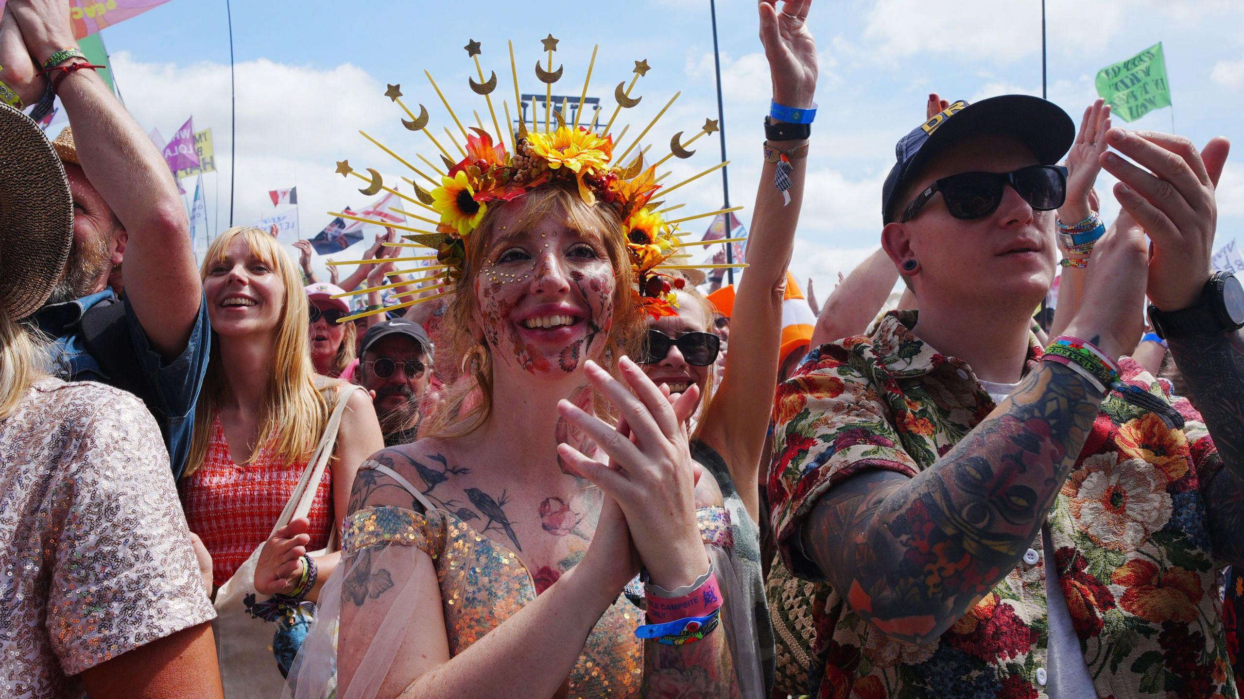 A woman wearing a flower crown and a glittery dress is clapping while standing in a crowd in front of the Pyramid stage.