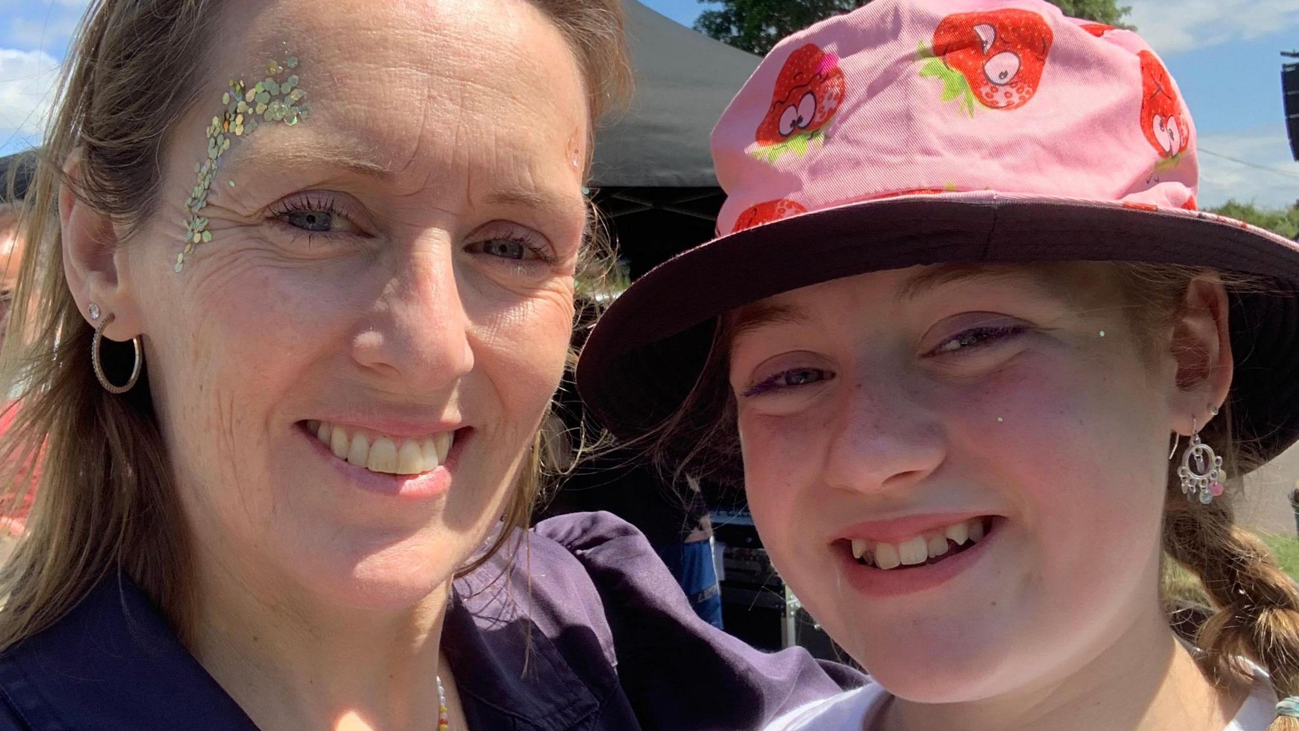 Jo on the left with glitter on her face and smiling, with her arm wrapped around Stella who is wearing a pink hat with strawberries on with her hair in braids