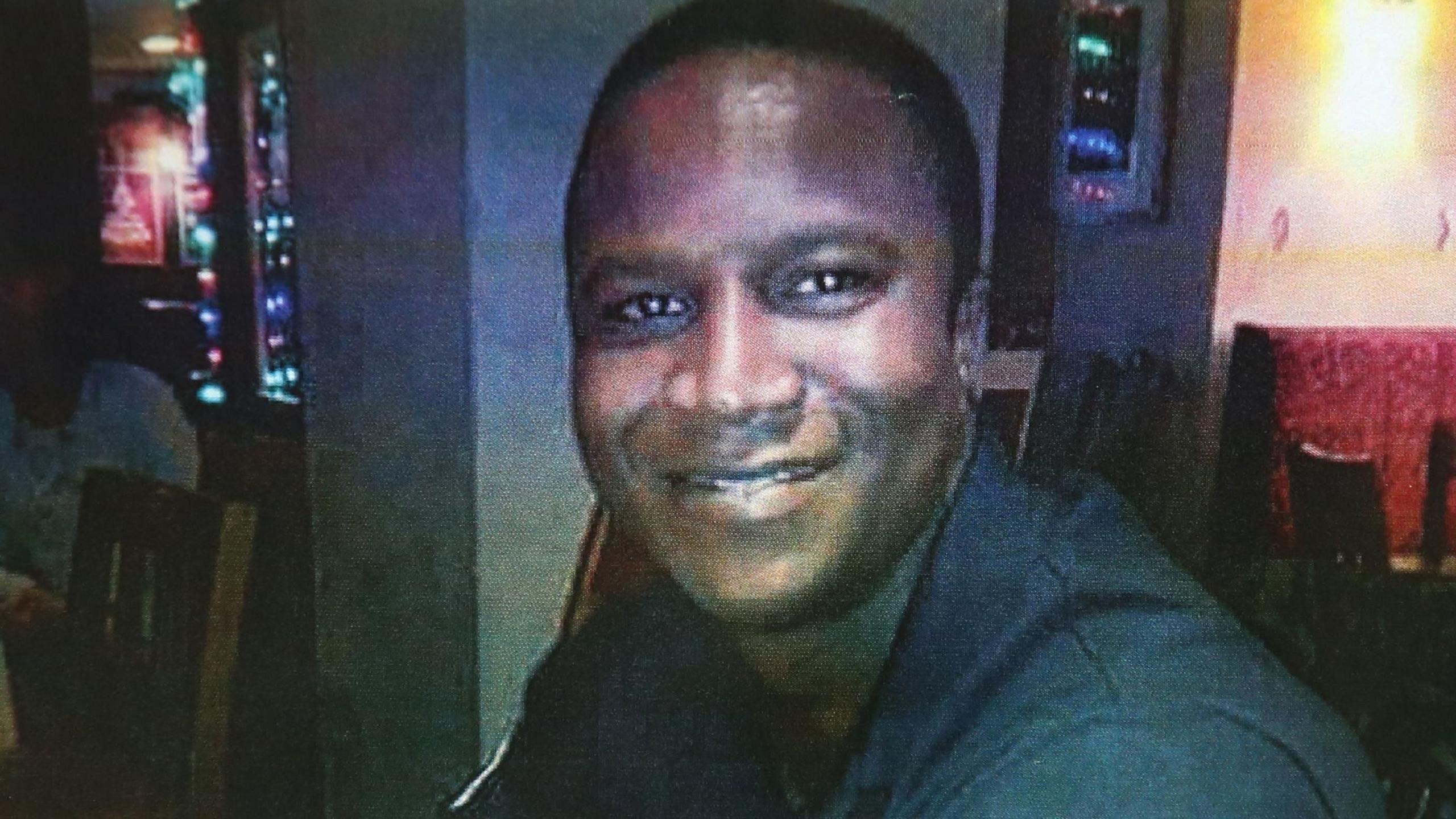 Sheku Bayoh is sitting in a bar in a dark shirt, smiling while looking at the camera.