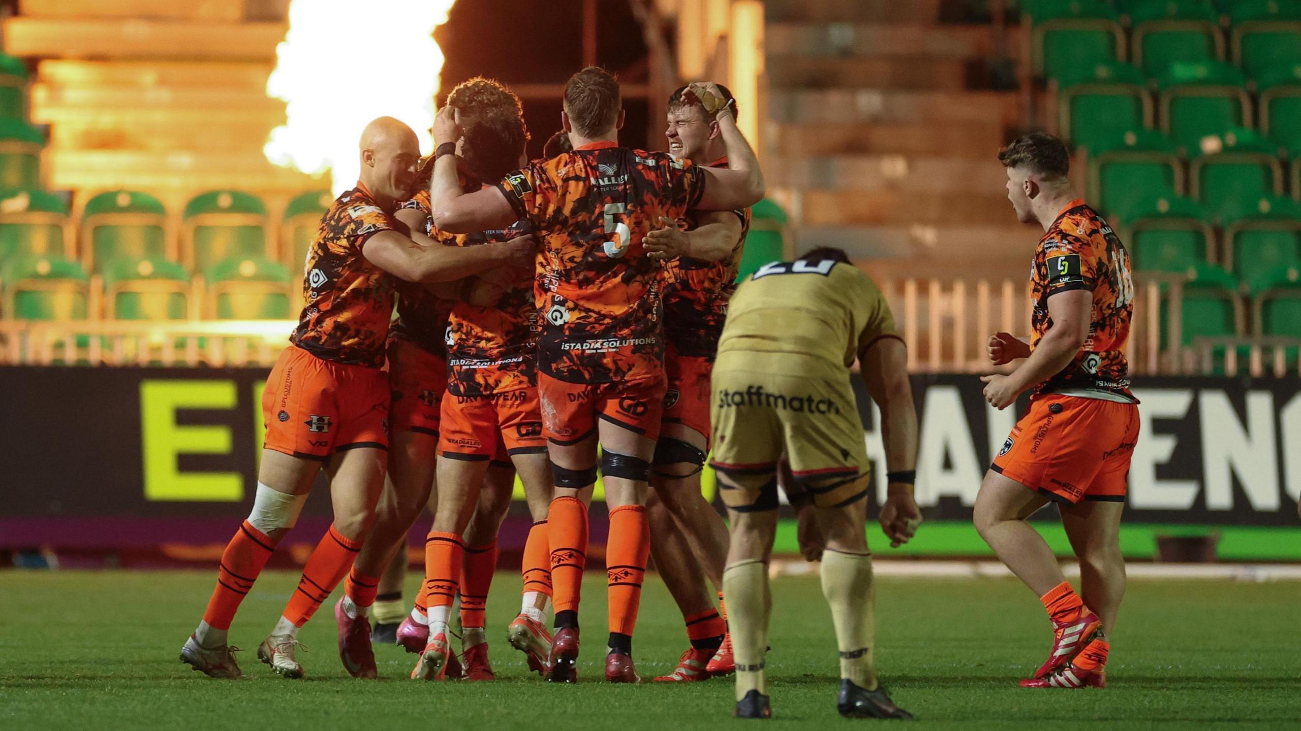 Dragons players in a huddle celebrating their victory against Lyon at Rodney Parade with a backdrop of empty seats and flamethrowers