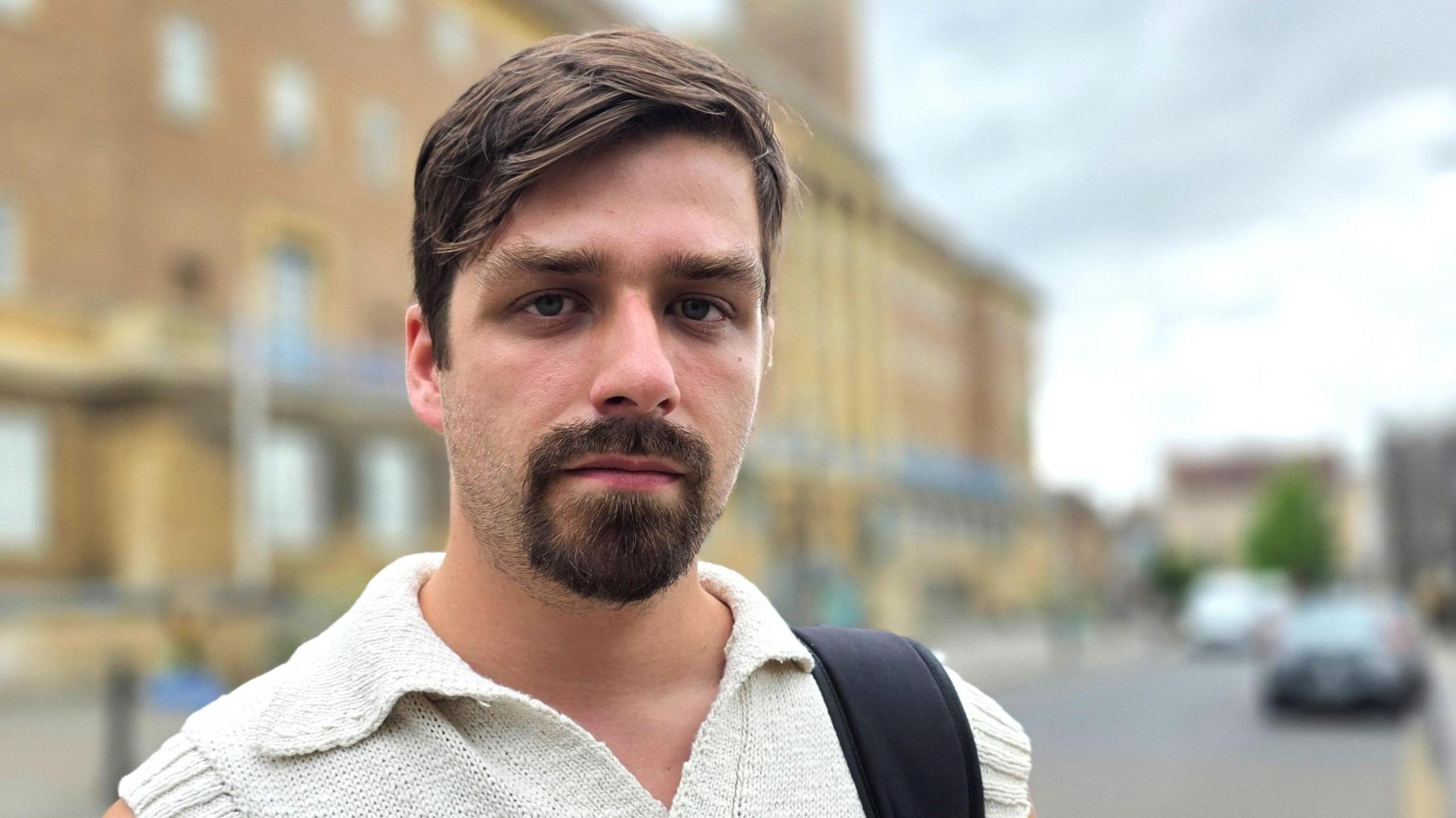 Alex Catt is wearing a short-sleeve, cream top. We can see a black bag strap over his left shoulder. Behind him is Norwich City Hall.