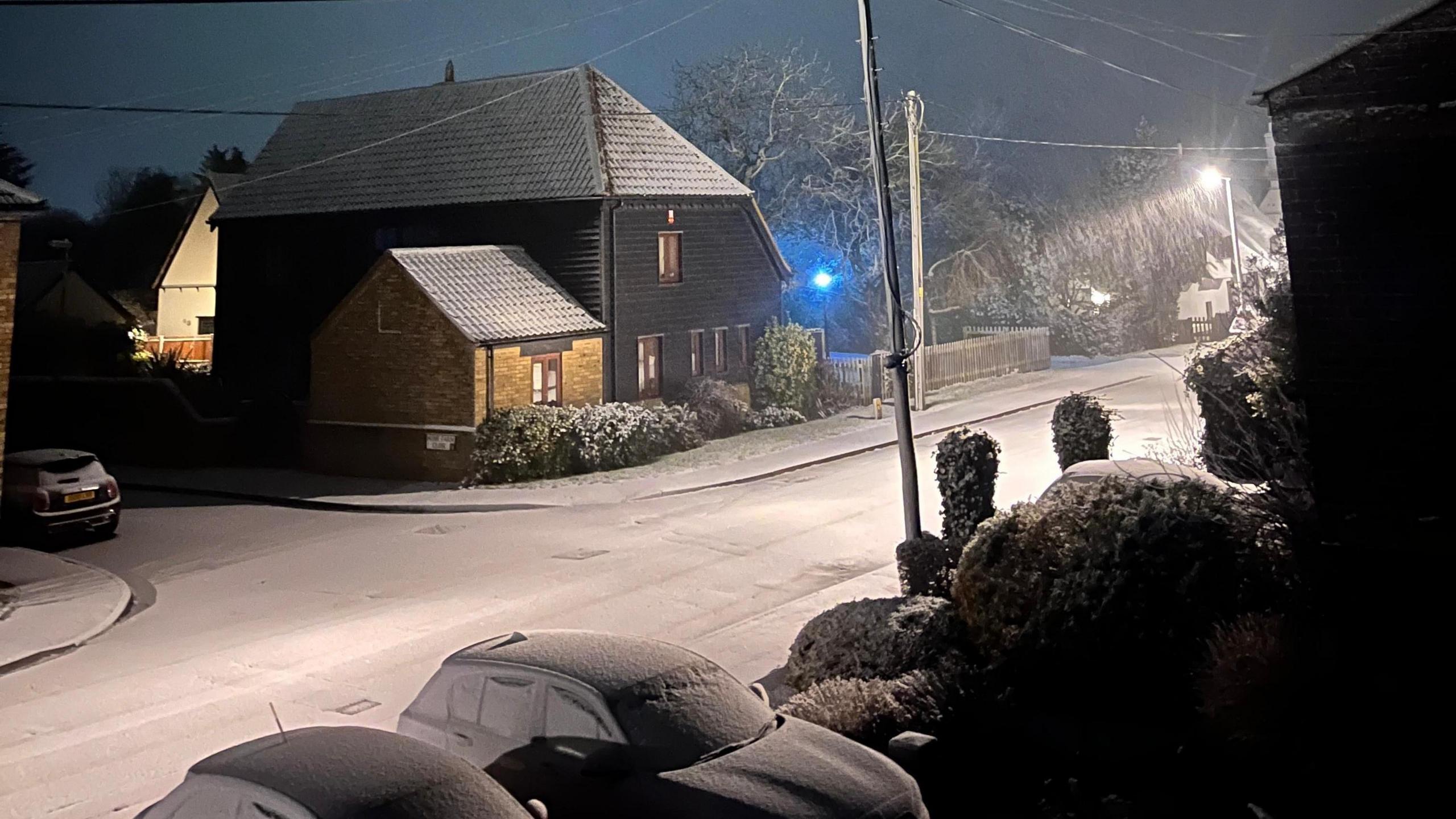 Snow on a road, showing snow on cars, trees and tops of houses. There is a street light on in the distance and it is dark. 