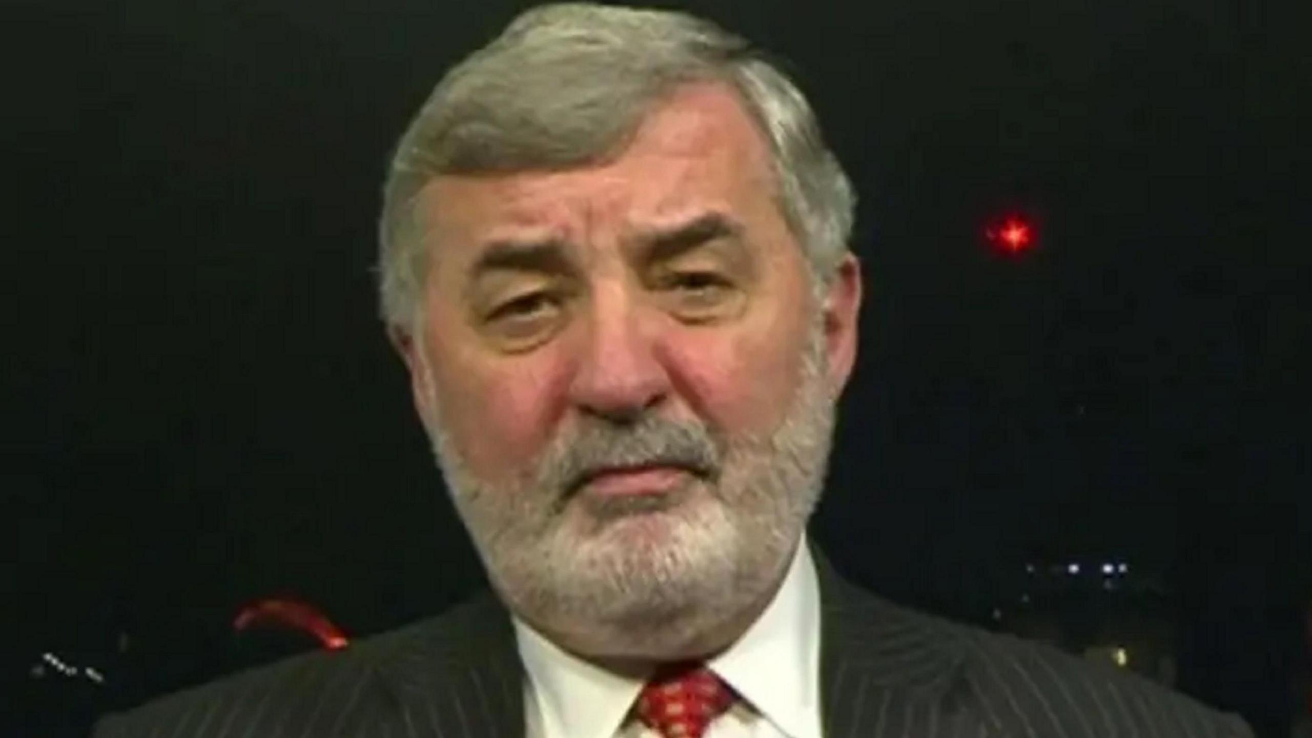 Lord Alderdice has grey hair parted to the side with a grey beard and is wearing a pin striped dark suit
