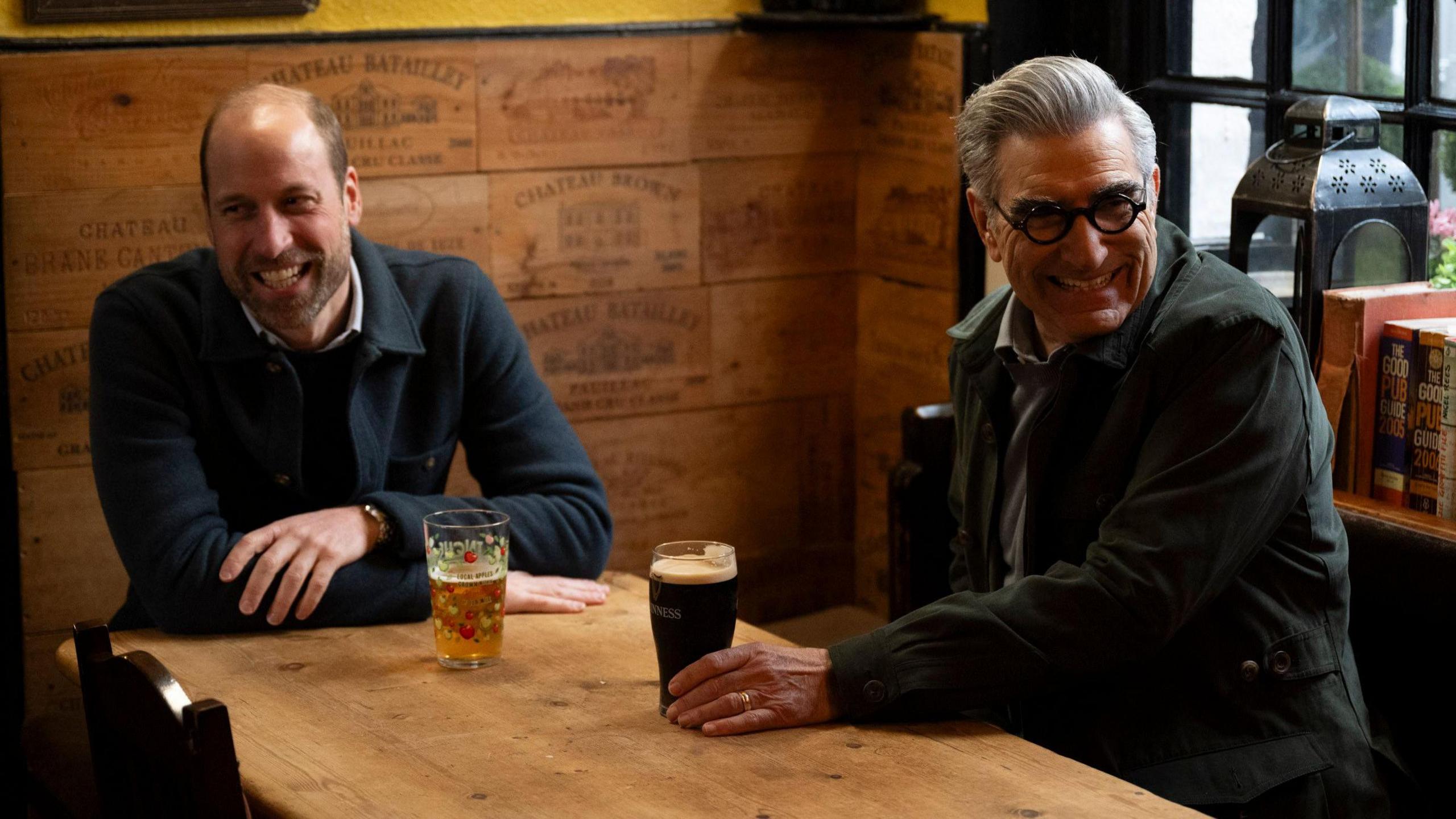 Prince William and Eugene Levy sharing a drink in a pub, both are smiling