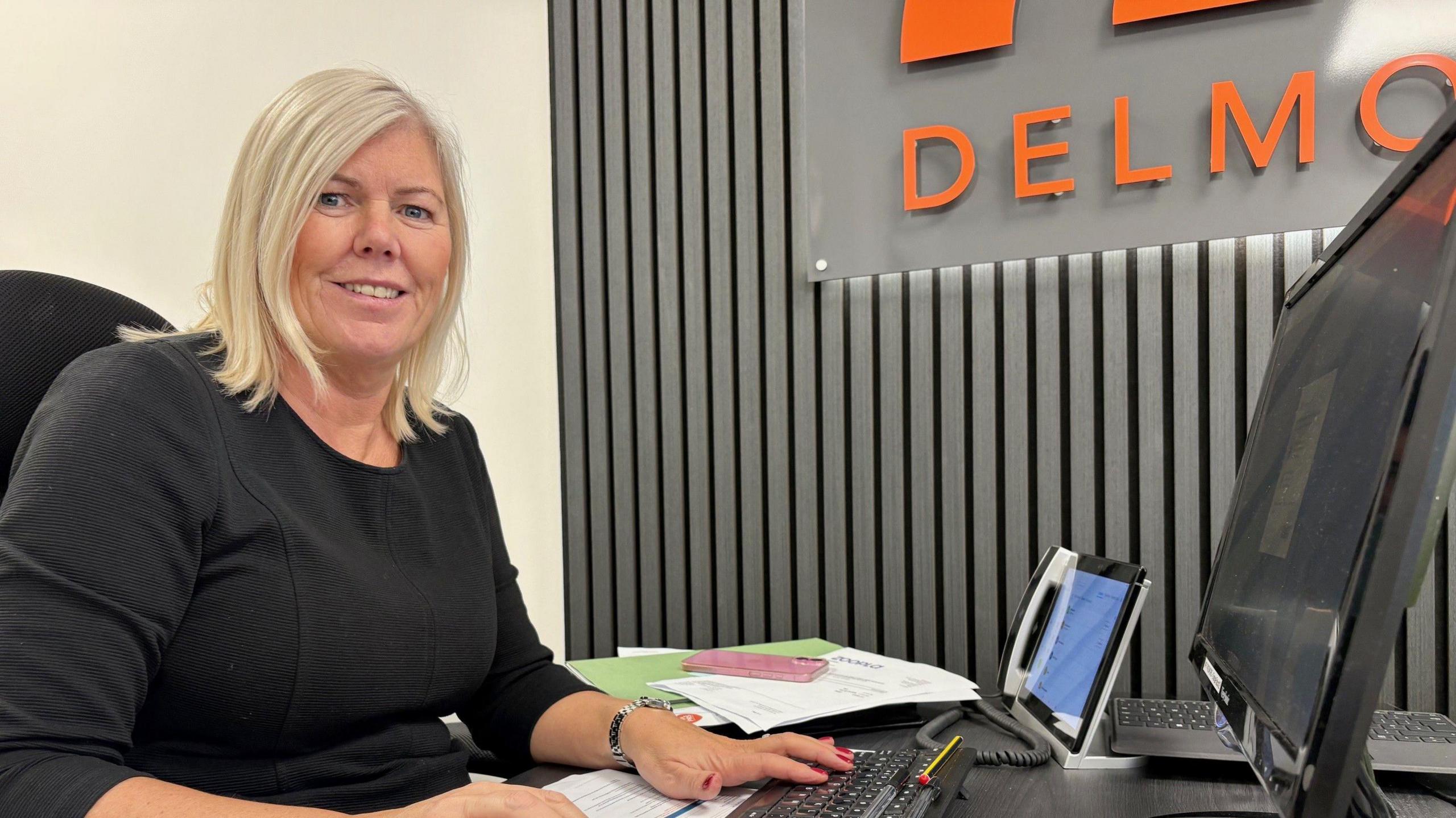 Angela has blonde hair and is wearing a black top. she is sitting at a computer screen.
