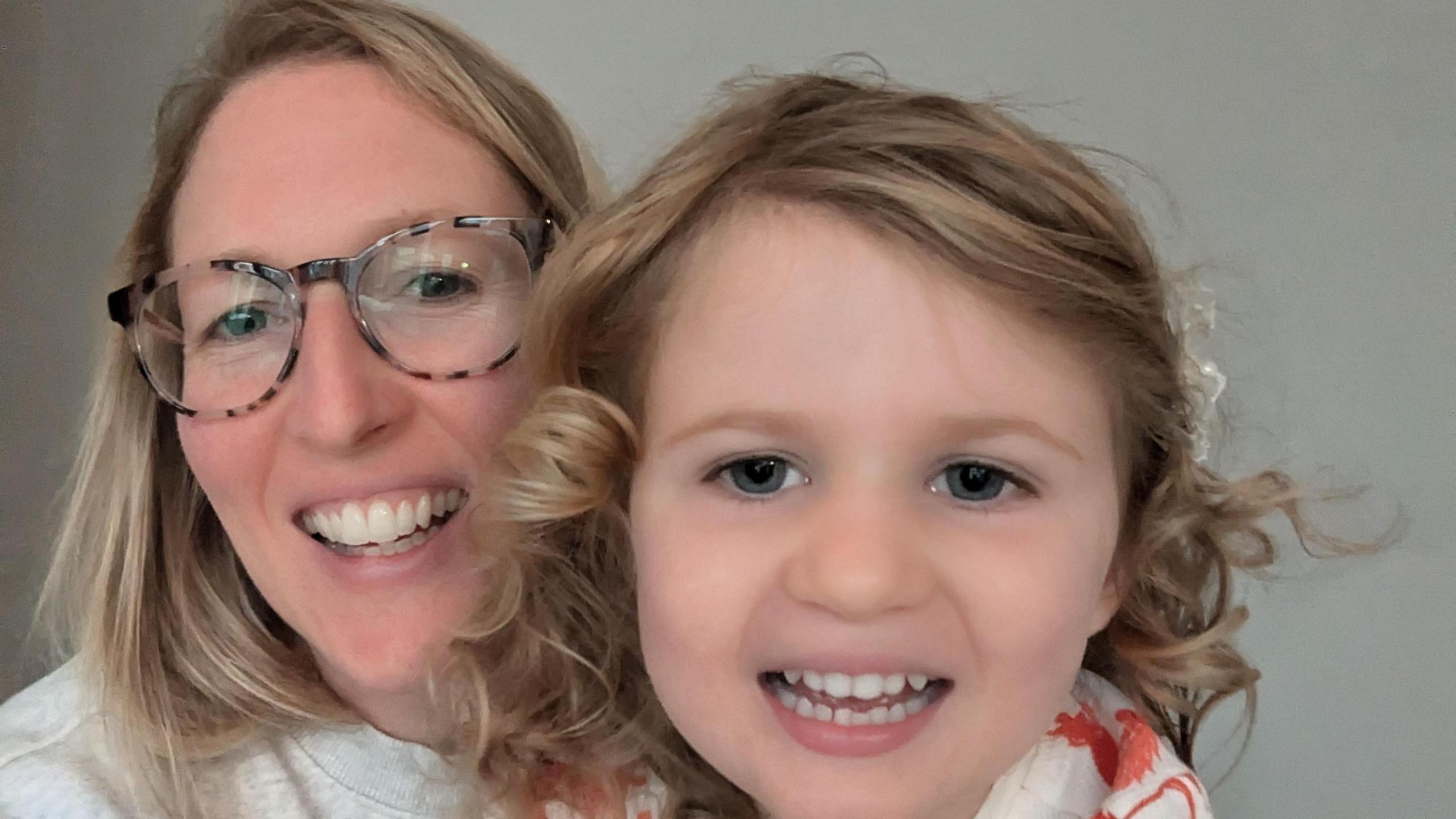 Megan, with blonde hair, glasses and a grey jumper, posing for a selfie with her daughter Joy, who has curly mousy blonde hair, is smiling and has big blue eyes