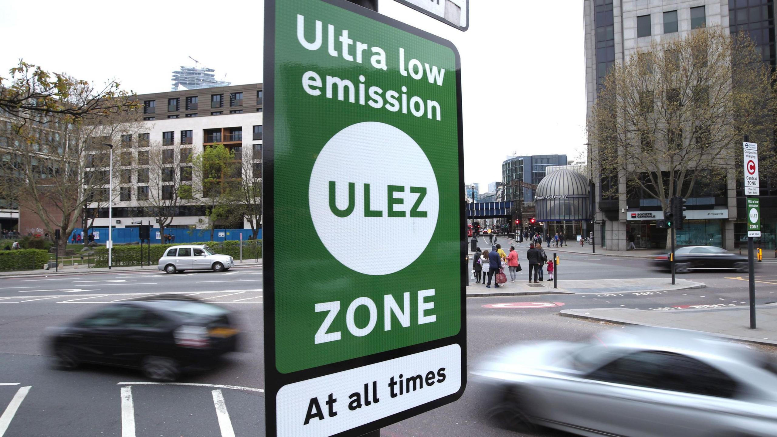 A sign saying 'ultra low emission zone - at all times' in green and white, at a road junction in near Tower Bridge with cars and buildings in the background.