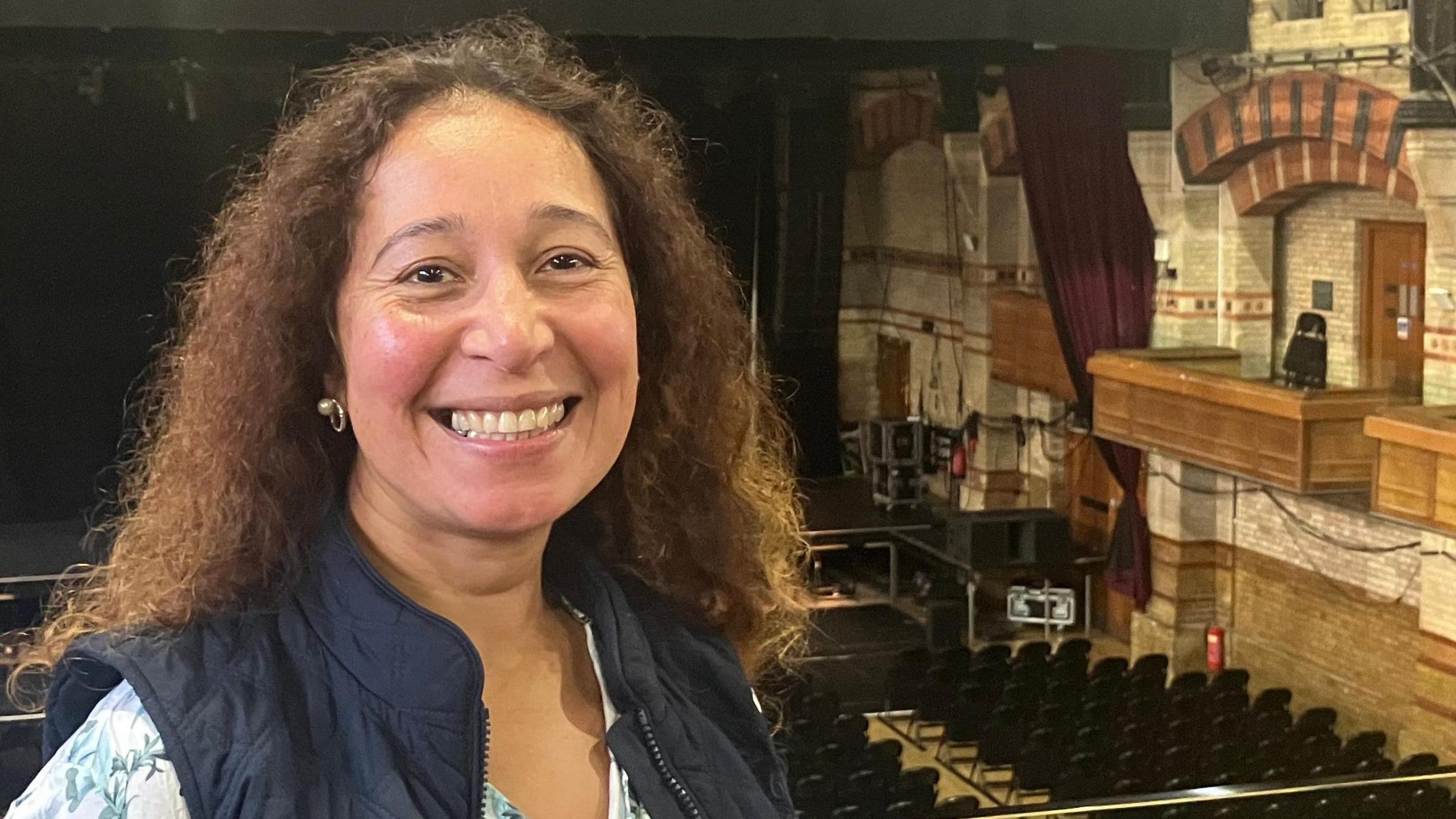 Antoinette Nestor stands inside Cambridge’s Corn Exchange. The warm lighting casts a soft glow across the historic brickwork, highlighting the venue’s ornate features. She wears a blue gilet over a crisp white blouse, her long curly brown hair is going over her shoulders. Behind her, the stage, chairs, and boxes of the Corn Exchange are visible.