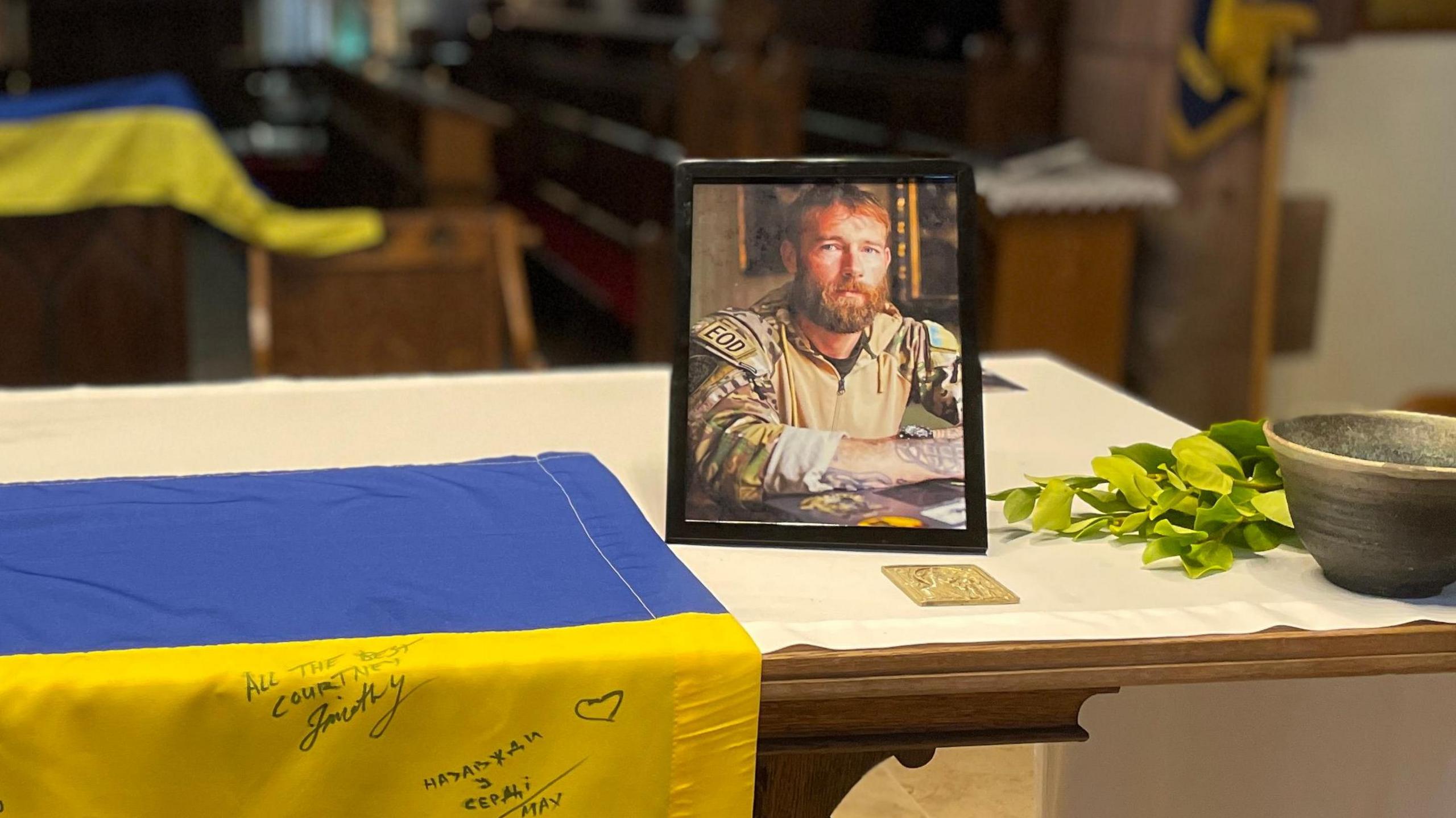 A framed phot of Chris Garrett in military fatigues placed on the alter with the blue and yellow of the Ukrainian flag in the background.