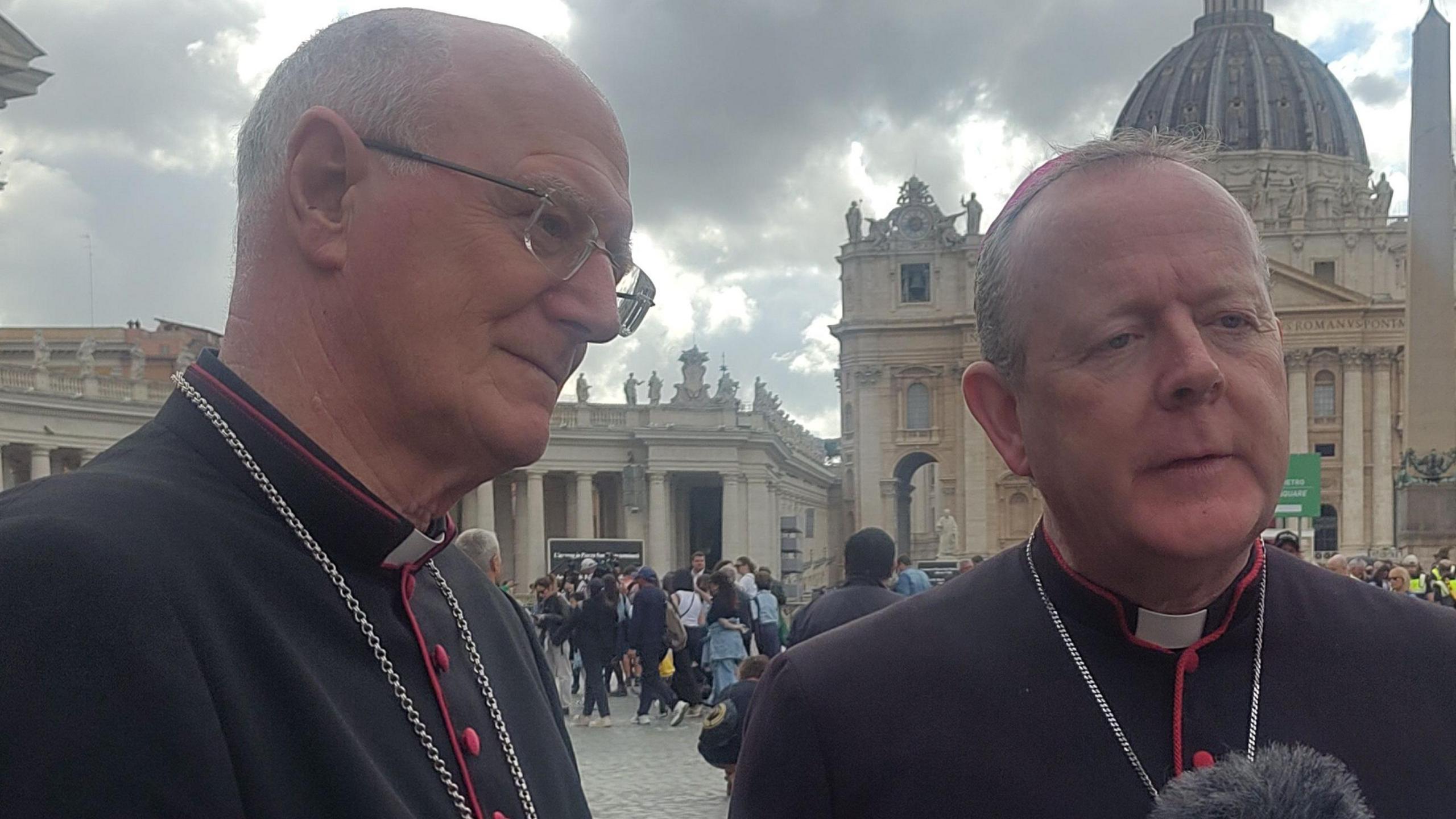 Dermot Farrell and Eamon Martin, who are both wearing traditional Catholic Church garments, speak to journalists in St Peter's Square. Behind them are tall, grand, light-coloured stone buildings.
