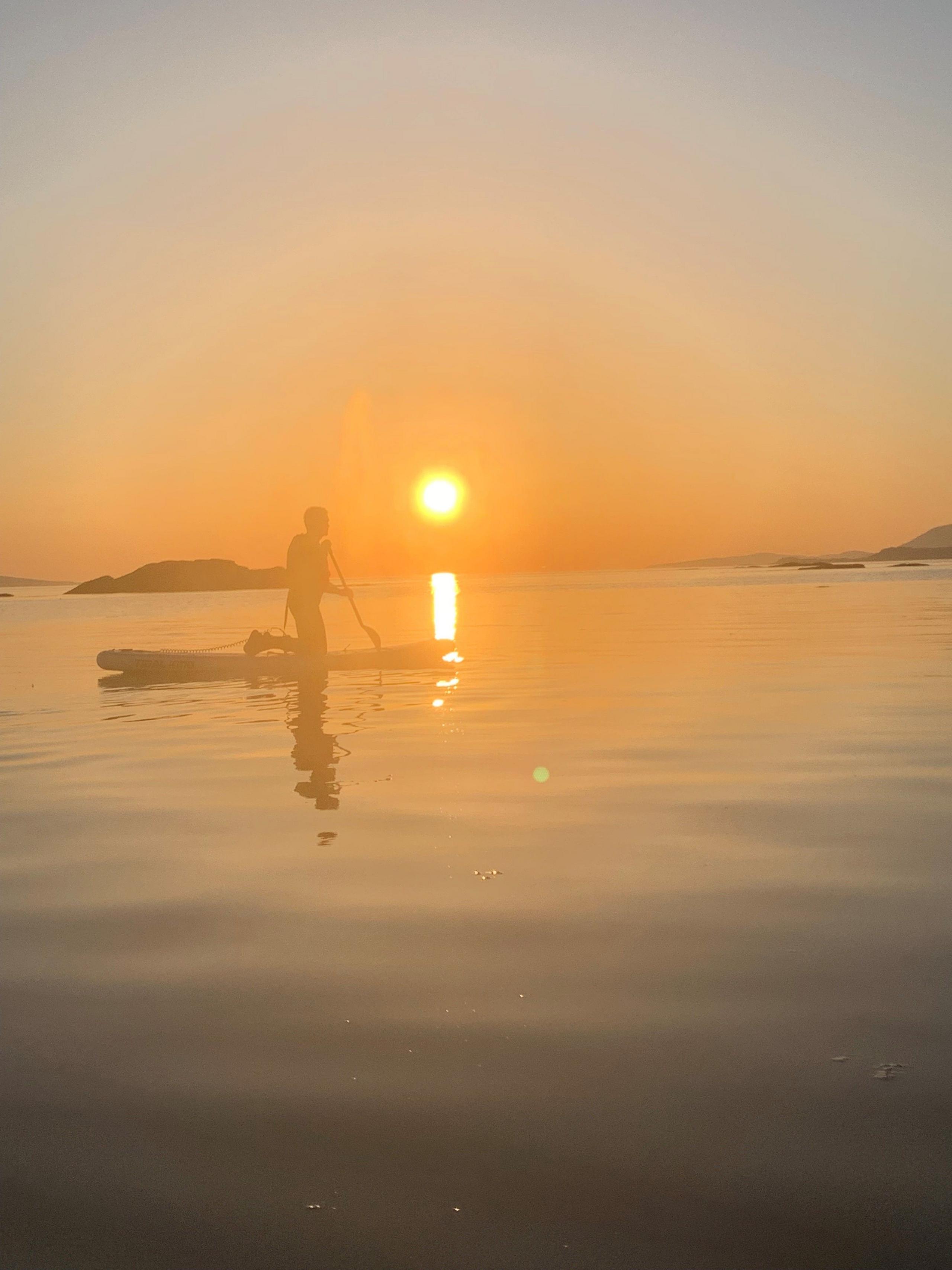 A paddle boarder goes across water with the sun setting, creating an orange hue in the sky