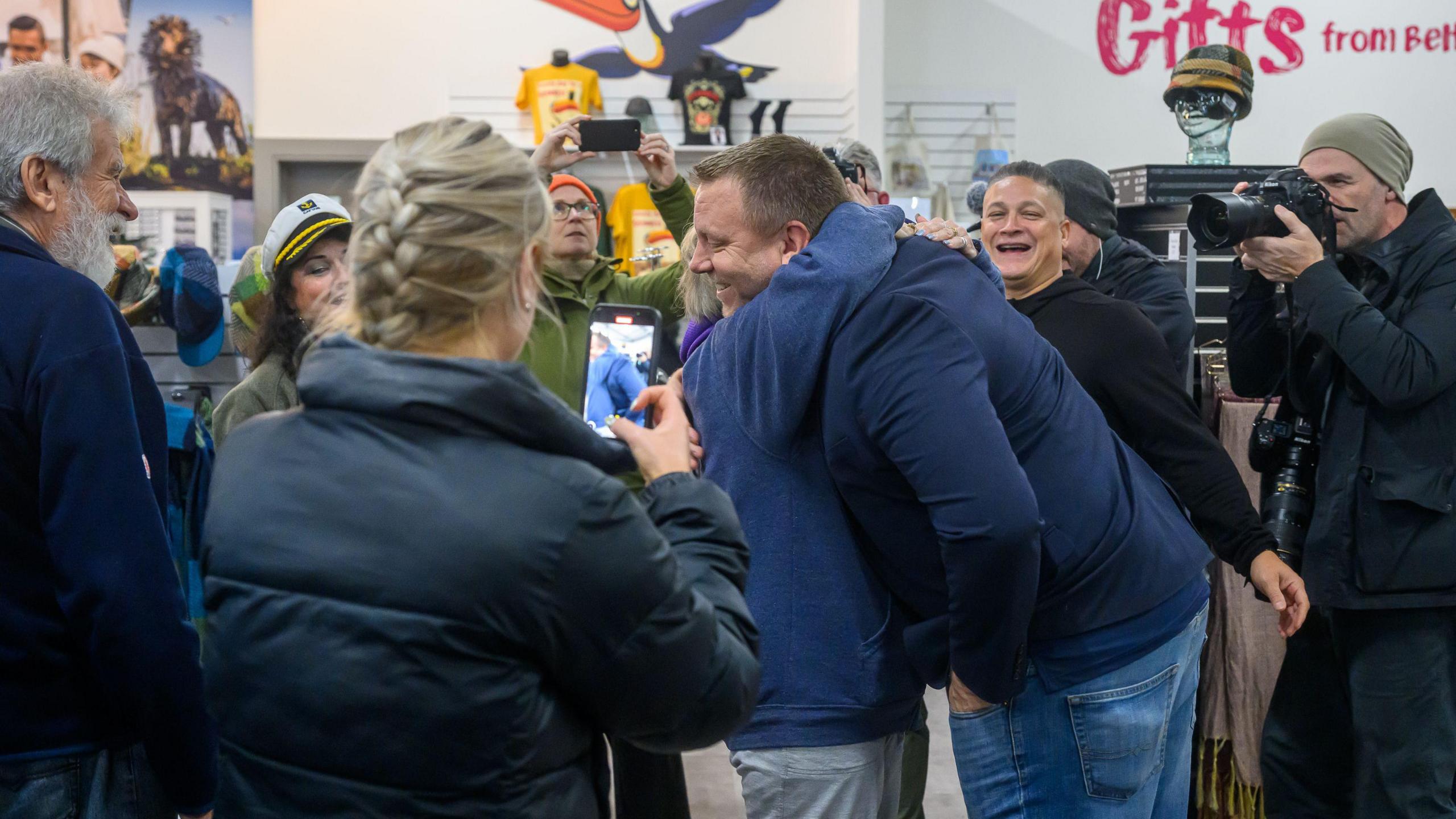 A woman hugging a man. They are surrounded by people smiling and taking photos and videos of them. 