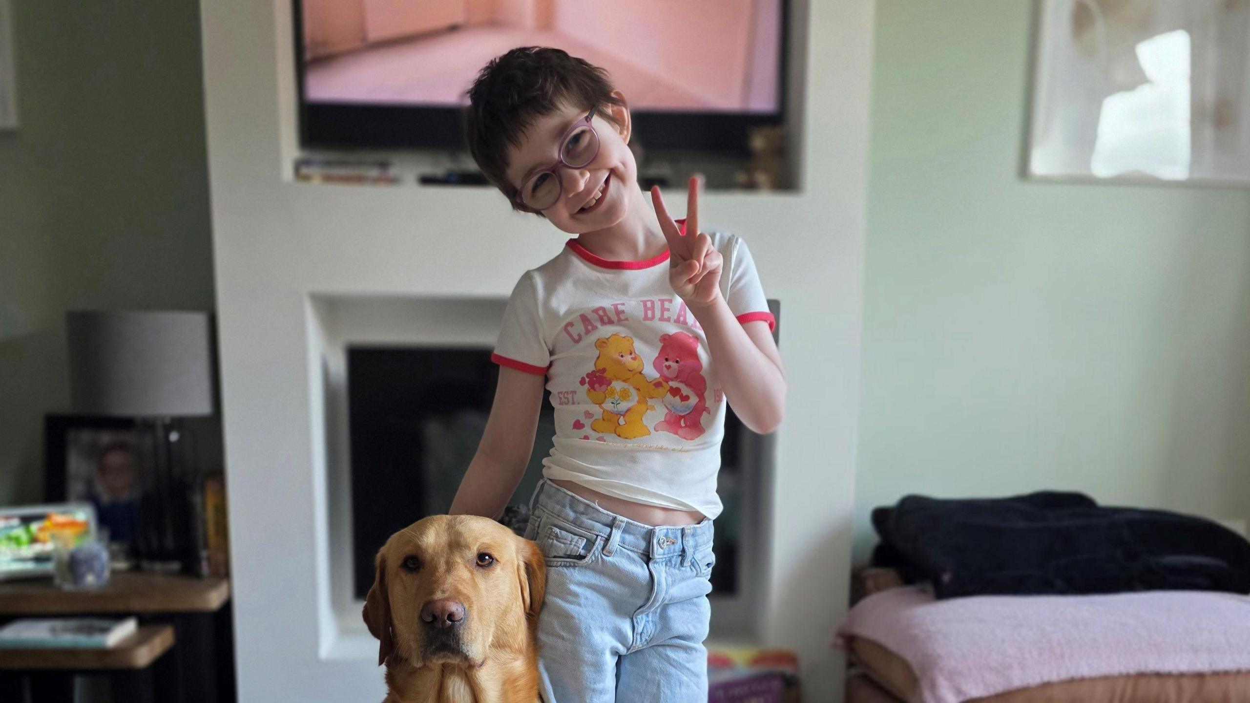 A young girl with short dark hair, purple-framed glasses, a white T-shirt with the words Care Bears on it, and pale blue jeans. She has her right hand on a light brown dog and her left is doing a V for victory symbol. She is standing in front of a fireplace with a TV screen above it and there is a chair with a pink cushion on it to the side.