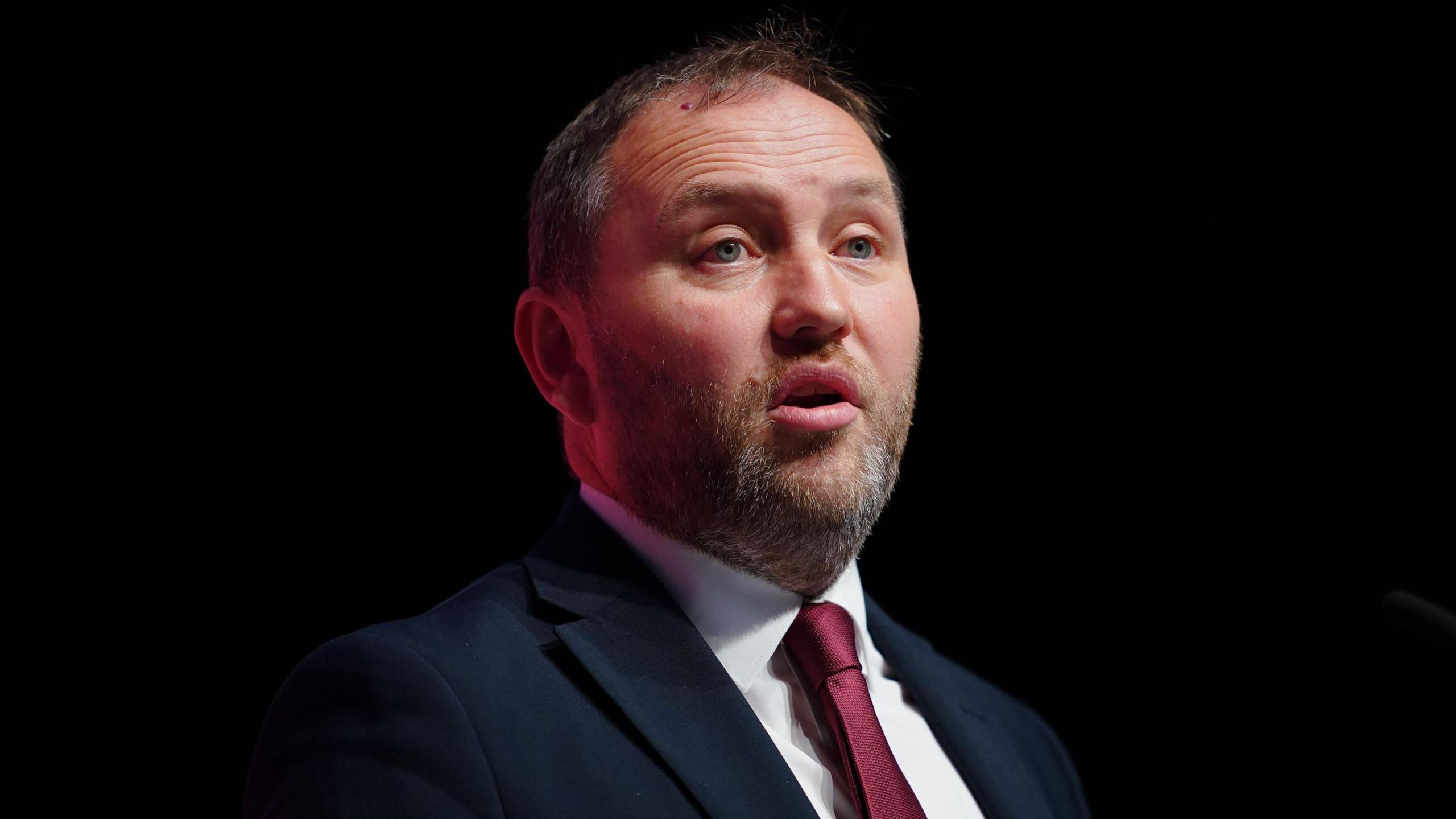 Ian Murray looking slightly off camera. He is only visible from the shoulders up. He is standing against a black background. He has short, dark hair and a grey and dark beard. His mouth is slightly open. He is wearing a dark suit over a white shirt with a dark red tie.