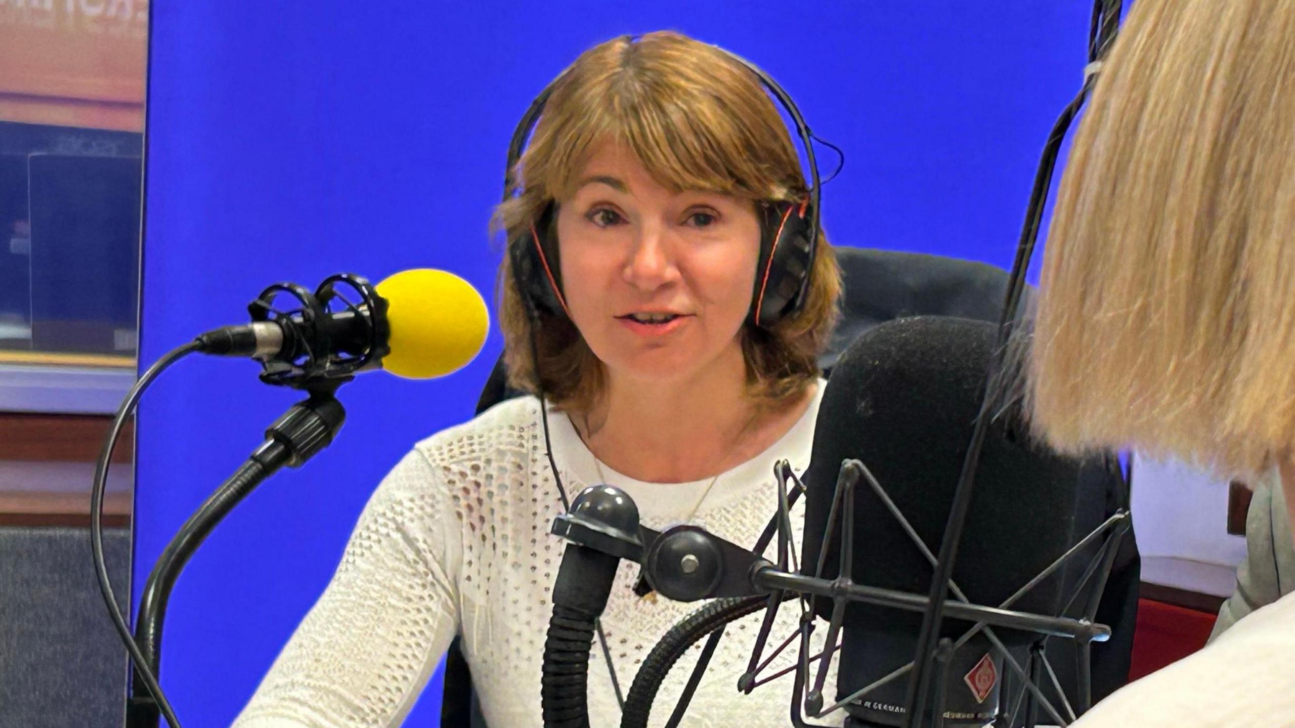 Lisa Spivey, wearing a white jumper, talks into a yellow microphone in the Radio Gloucestershire studio.