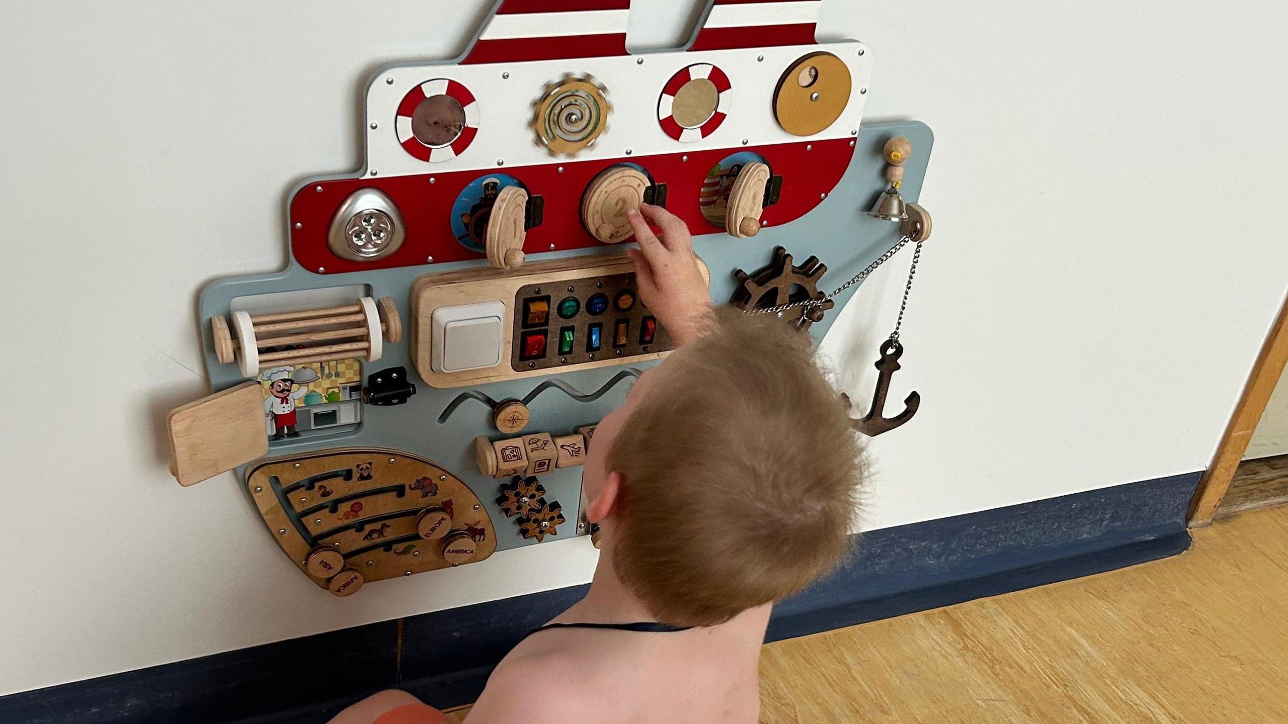 Connor wearing comfy orange shorts and grey socks with a green dinosaur on them. He is sitting on the floor of a hospital ward, playing with an interactive wooden ship on the wall, spinning the cogs and lifting handles.