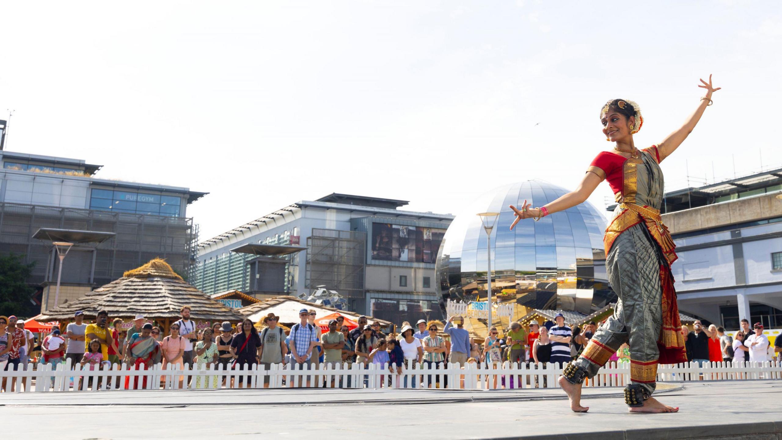 A woman in traditional Indian clothing, which is red, gold and silver, dancing in front of the Planetarium in Millennium Square