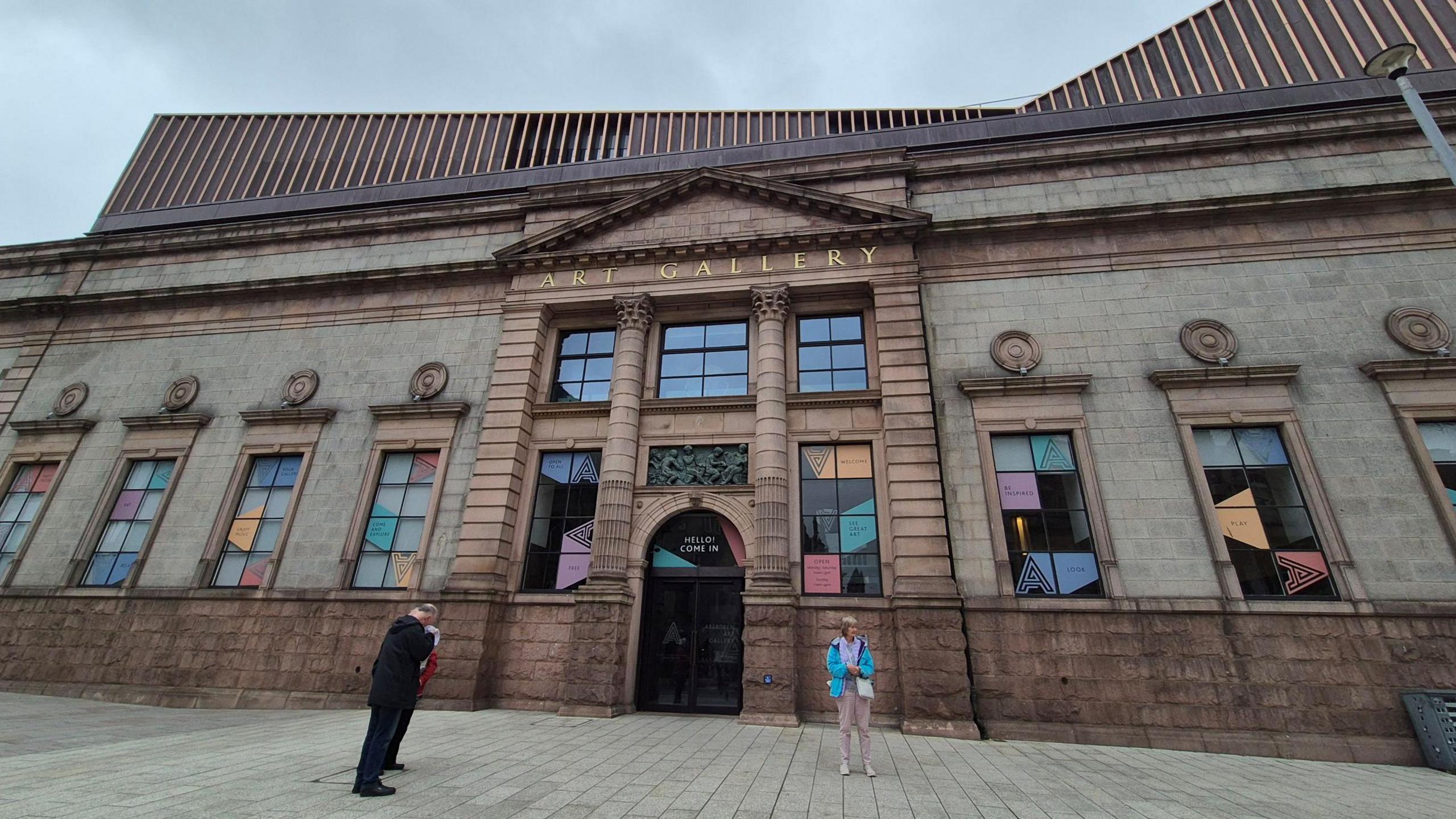 An exterior view of the front of Aberdeen Art Gallery. A stone building with columns and carvings, under a partly cloudy blue sky. The words “Art Gallery” are engraved in gold letters above the main entrance. A man and a woman are standing outside the building.