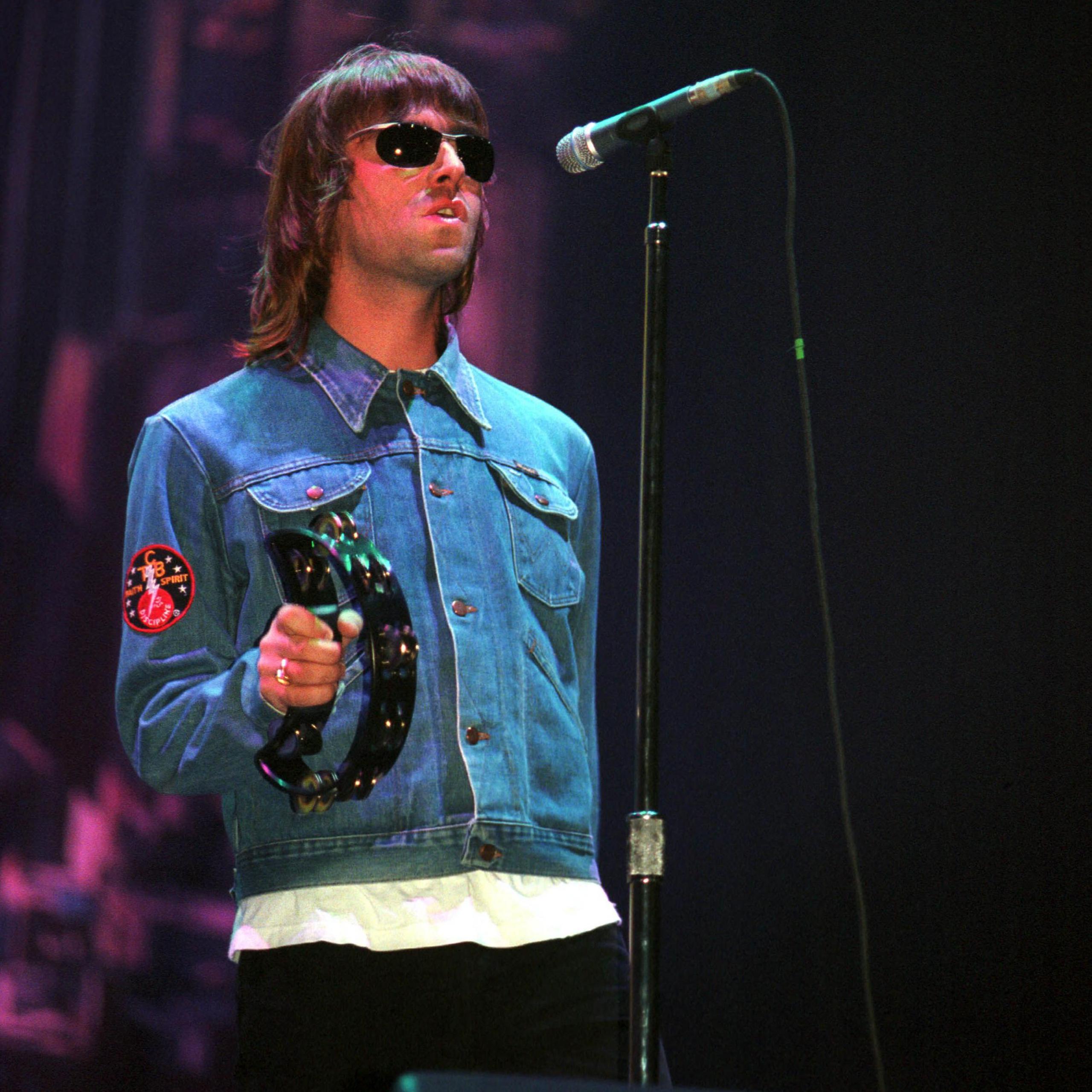 Liam Gallagher performing at Glasgow Green, on stage wearing a blue denim jacket holding a tamborine