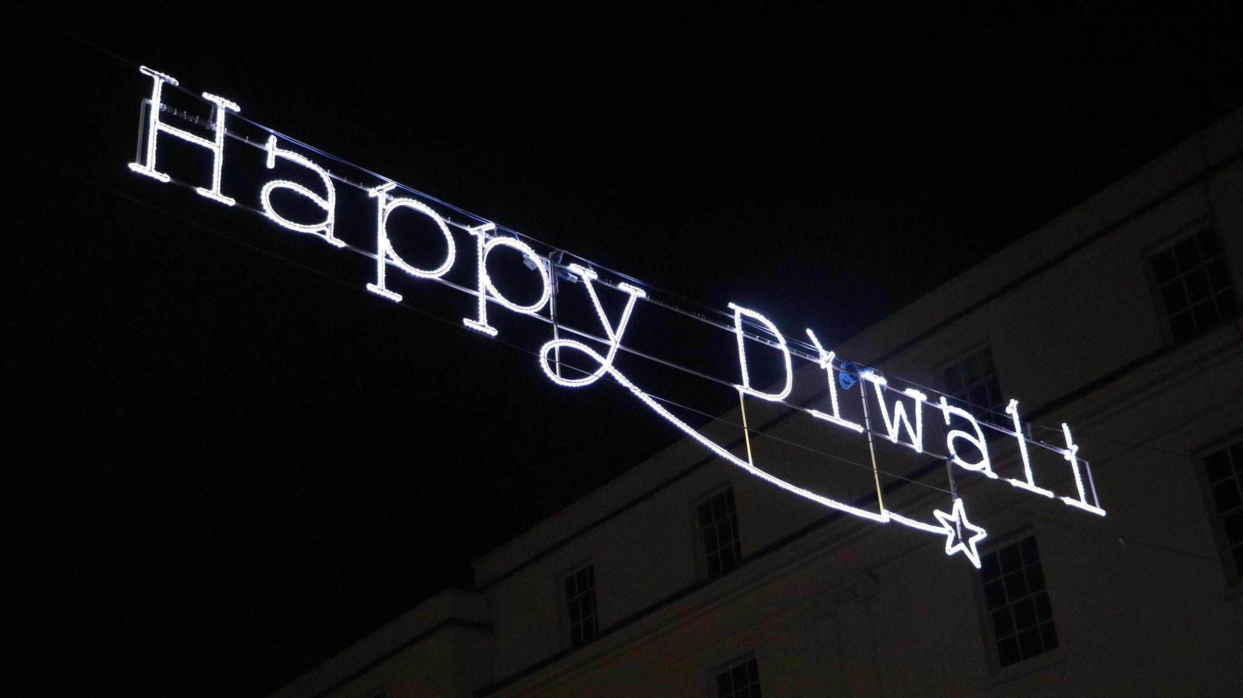 A neon light hangs between buildings, with the text 'Happy Diwali'.